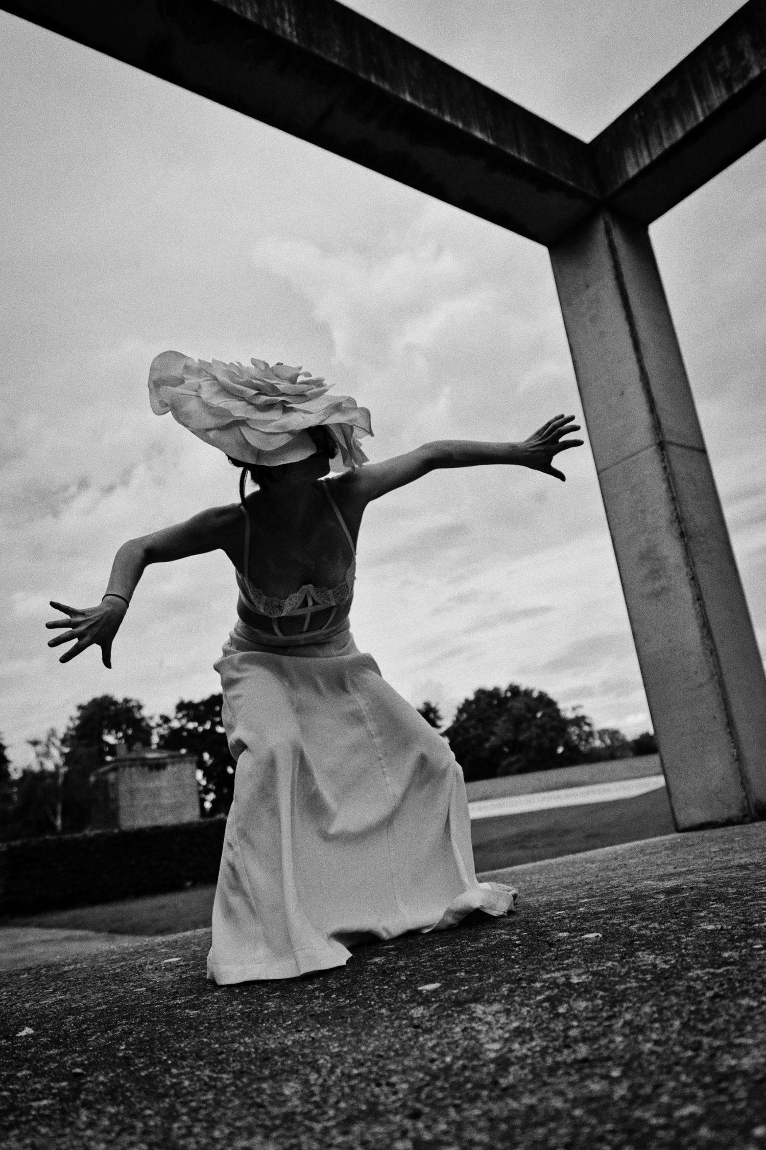 Black and white photo of a person in a flowing skirt and wide-brimmed hat dancing with outstretched arms beneath a concrete archway.