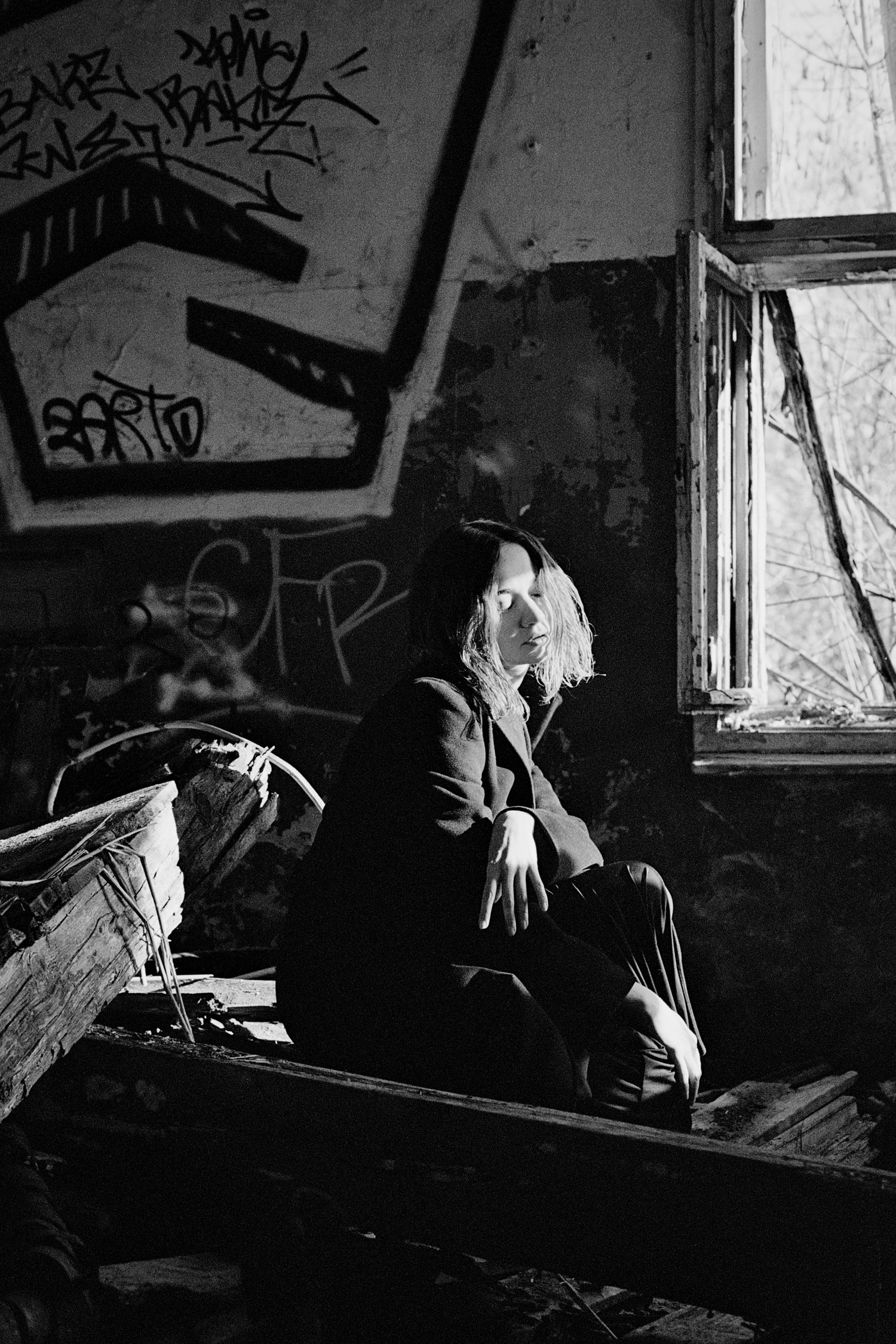 Black and white photo of a person sitting pensively in an abandoned building with graffiti-covered walls and broken windows.