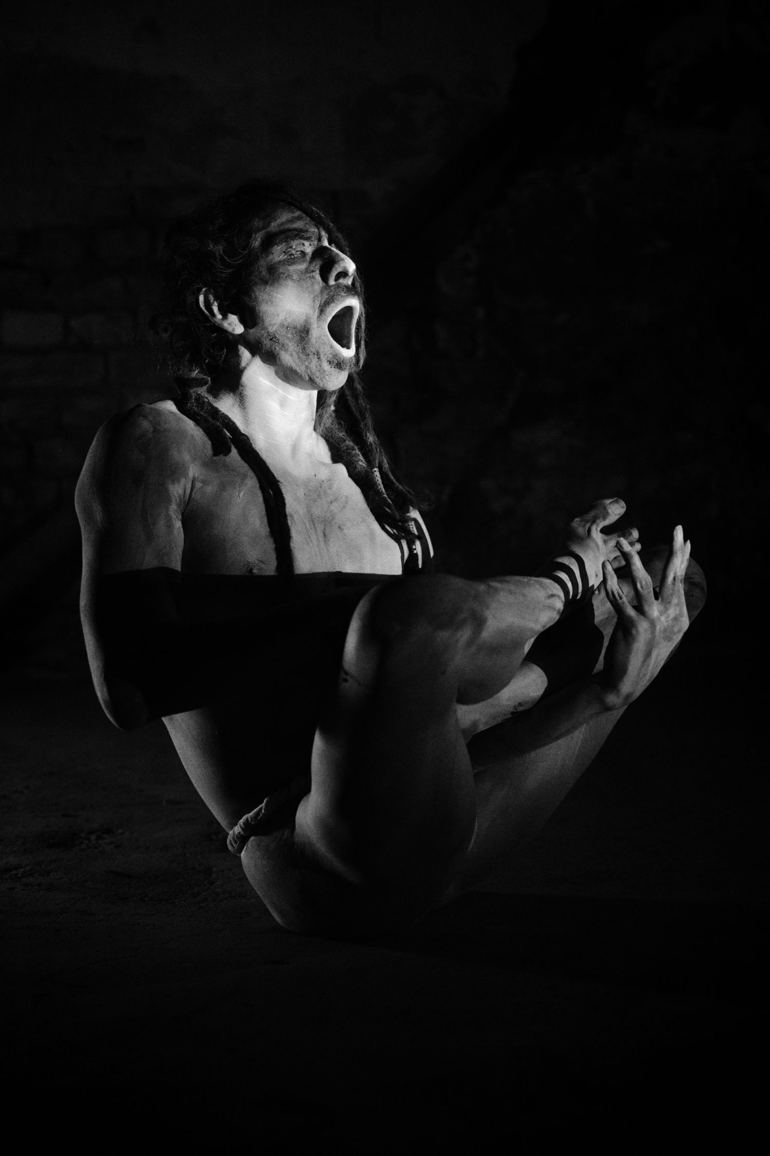 Black and white photo of a shirtless person kneeling, screaming dramatically with hands clasped in an artistic pose.