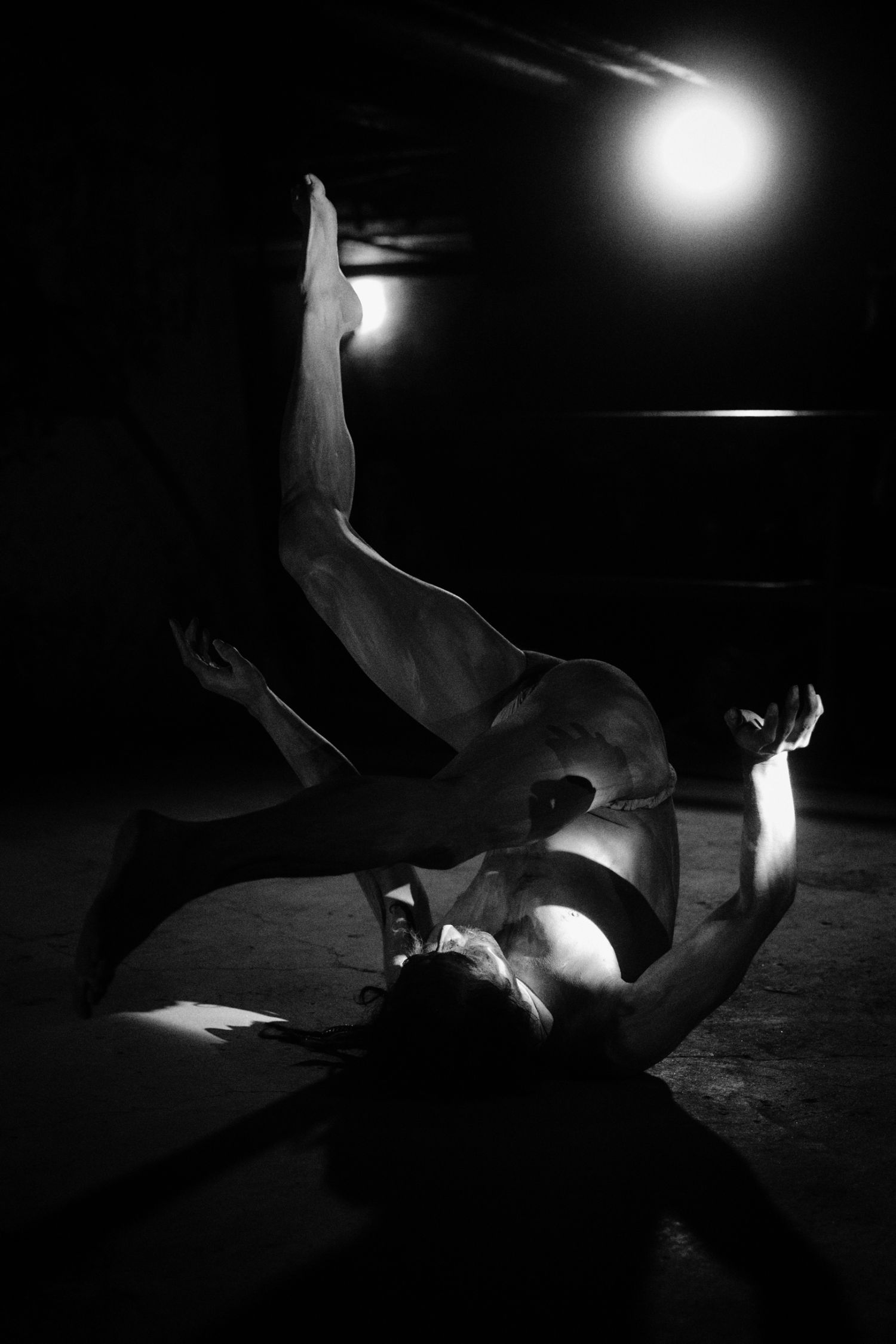 Black and white photo of a dancer performing a dramatic floor pose under a single spotlight in a dark studio.