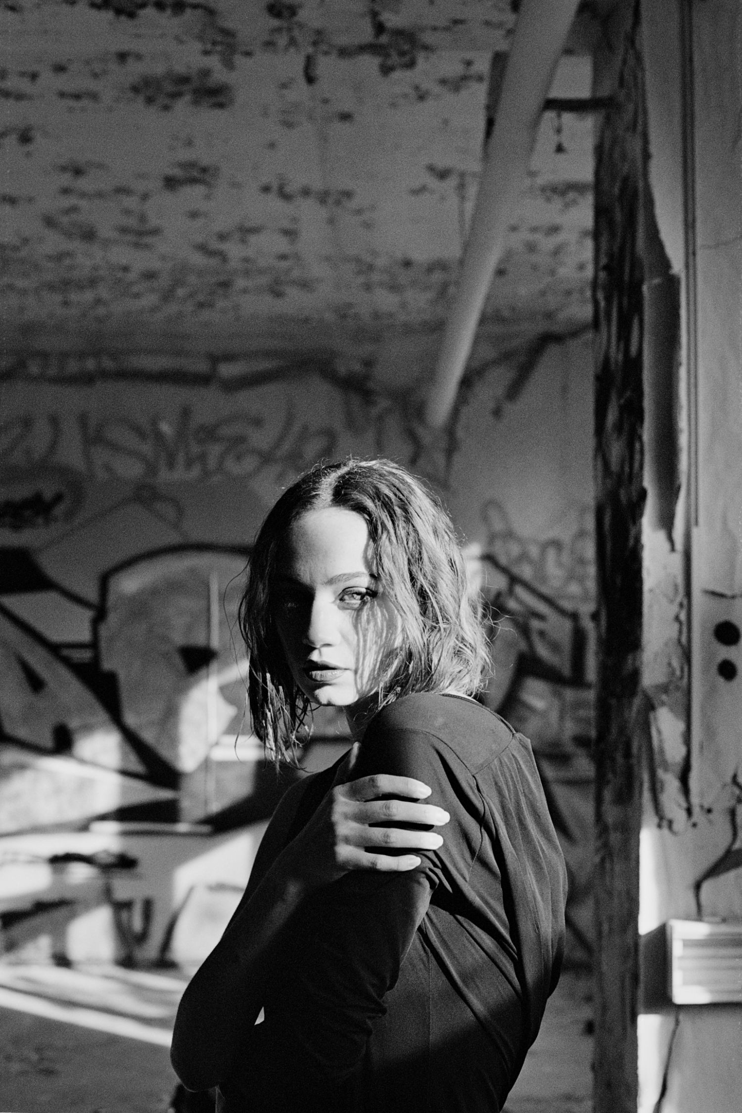 Black and white portrait of a young woman in dark clothing, standing in an abandoned building with graffiti walls.