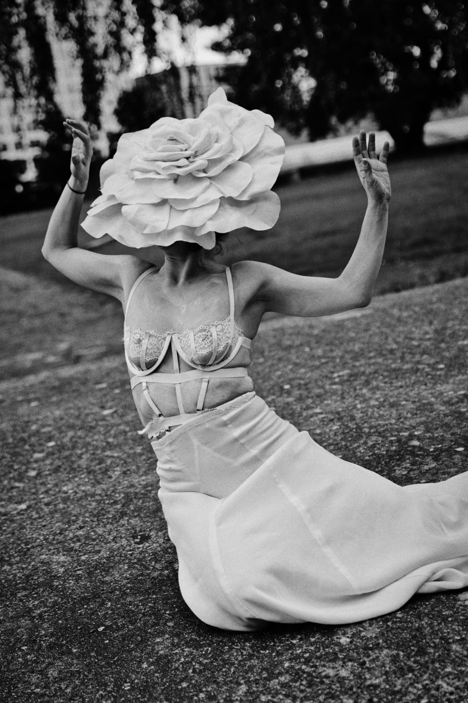 Black and white photo of a woman kneeling outdoors in a lace corset and flowing skirt, her face hidden by an oversized flower hat.