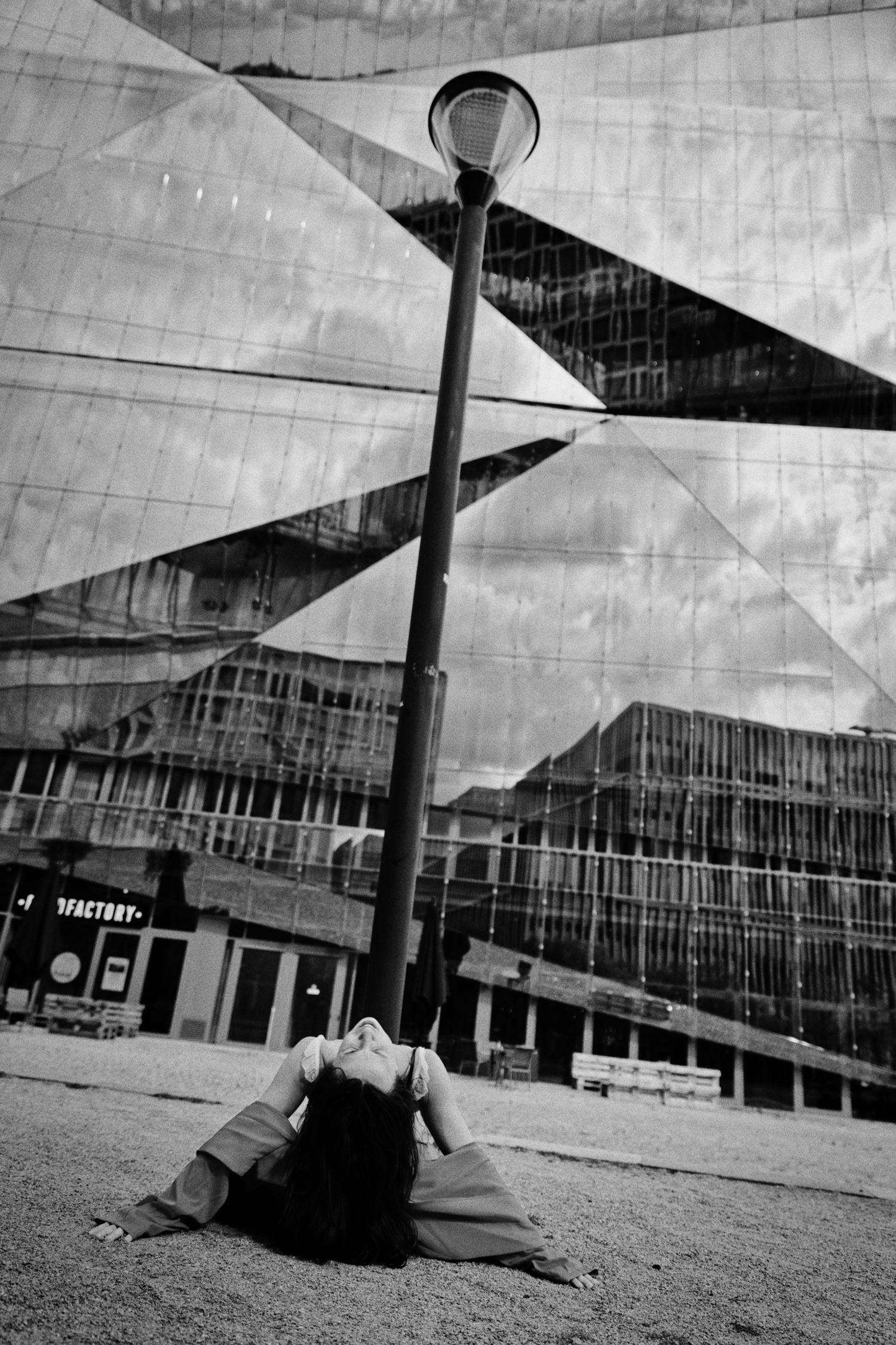 Black and white photo of a woman arching backward on the ground beside a street lamp, with a faceted mirrored glass building behind her.