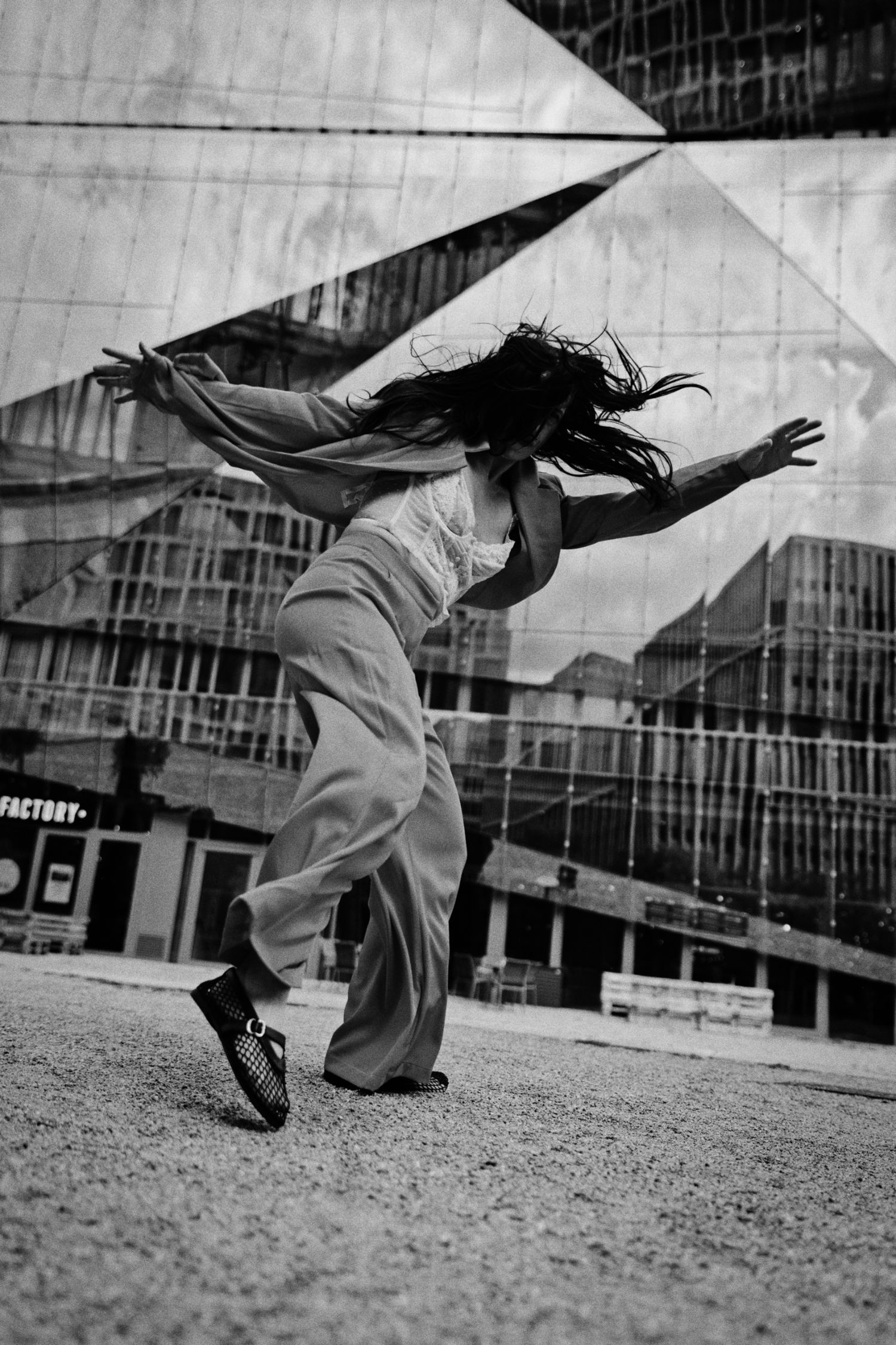 Black and white photo of a dancer in wide-leg trousers leaning with arms outstretched and hair flying before a mirrored glass building.