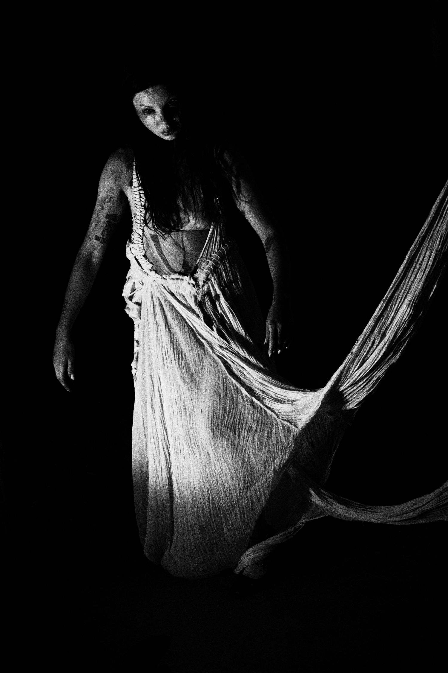 Black and white photo of a woman in flowing white dress emerging from darkness in dramatic artistic portrait.