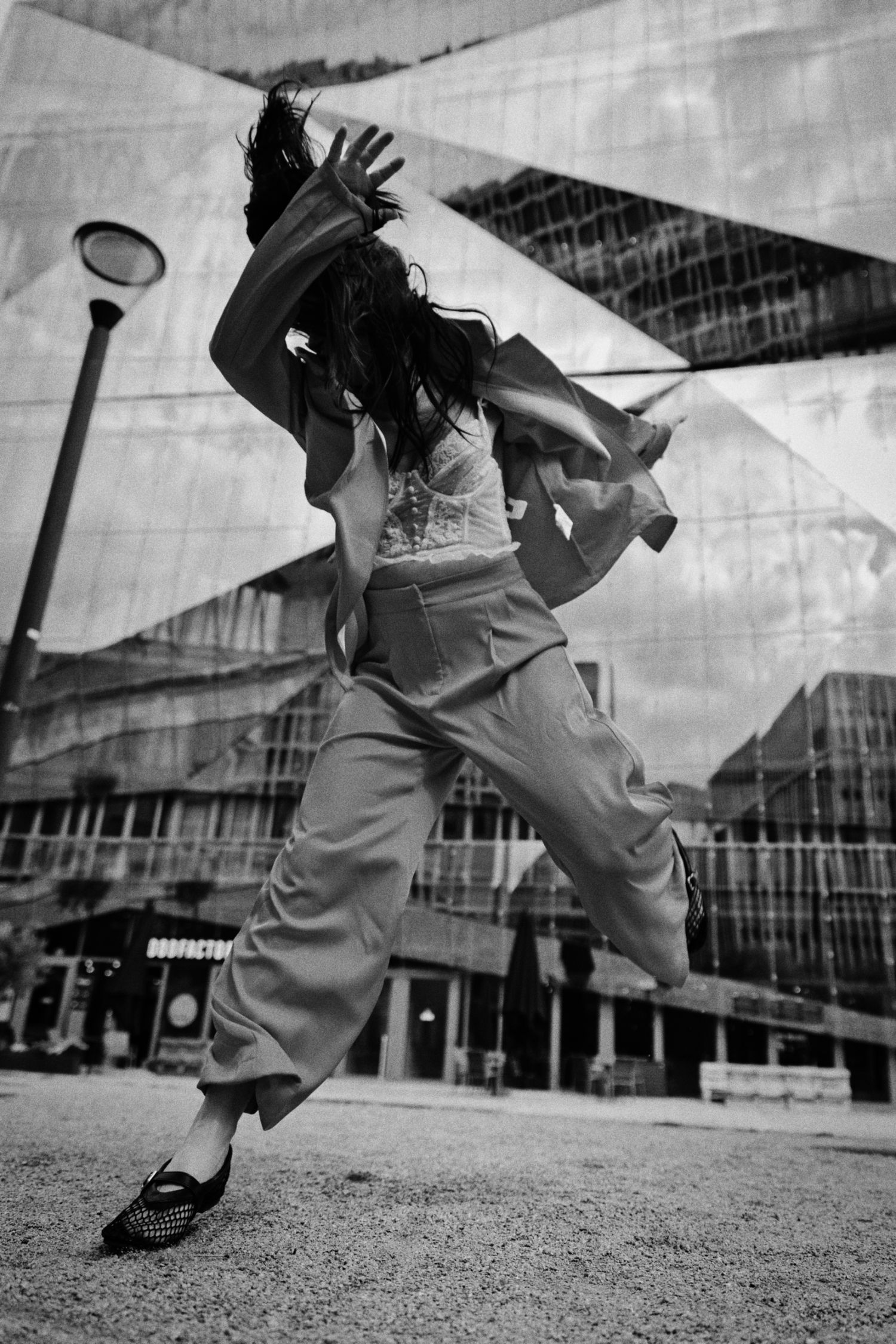 Black and white photo of a woman in wide-leg trousers and a loose jacket leaping mid-air in front of a modern glass building.