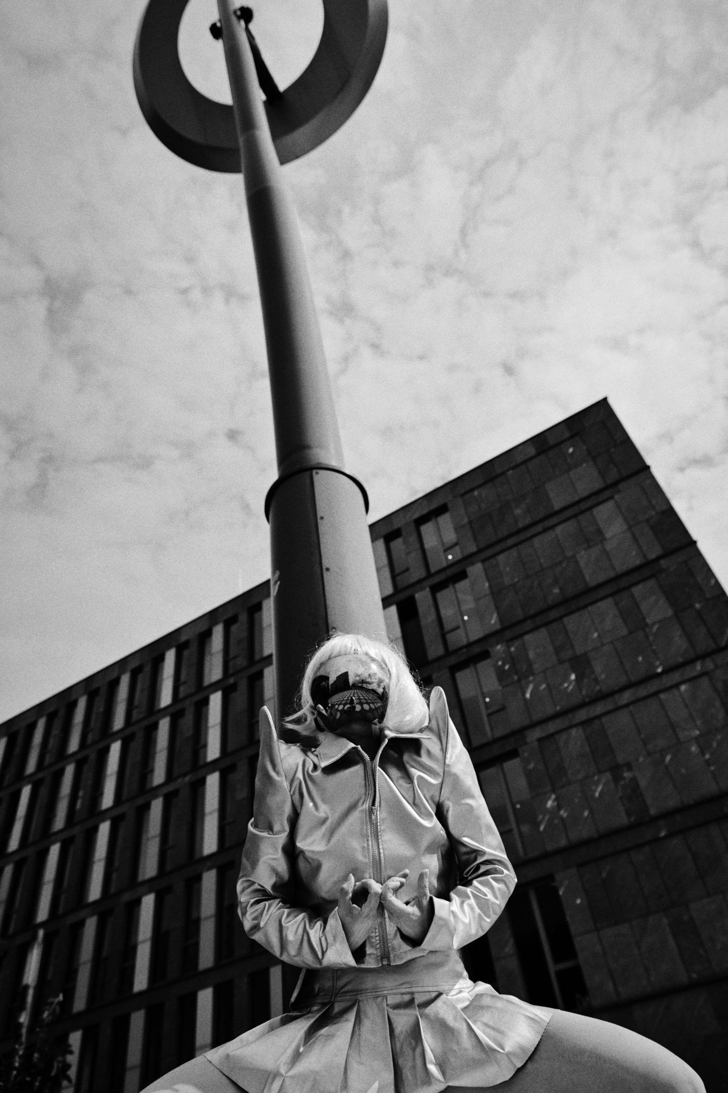 Black and white low-angle photo of a masked person in a light trench coat posing beneath a tall street lamp near a modern glass building.