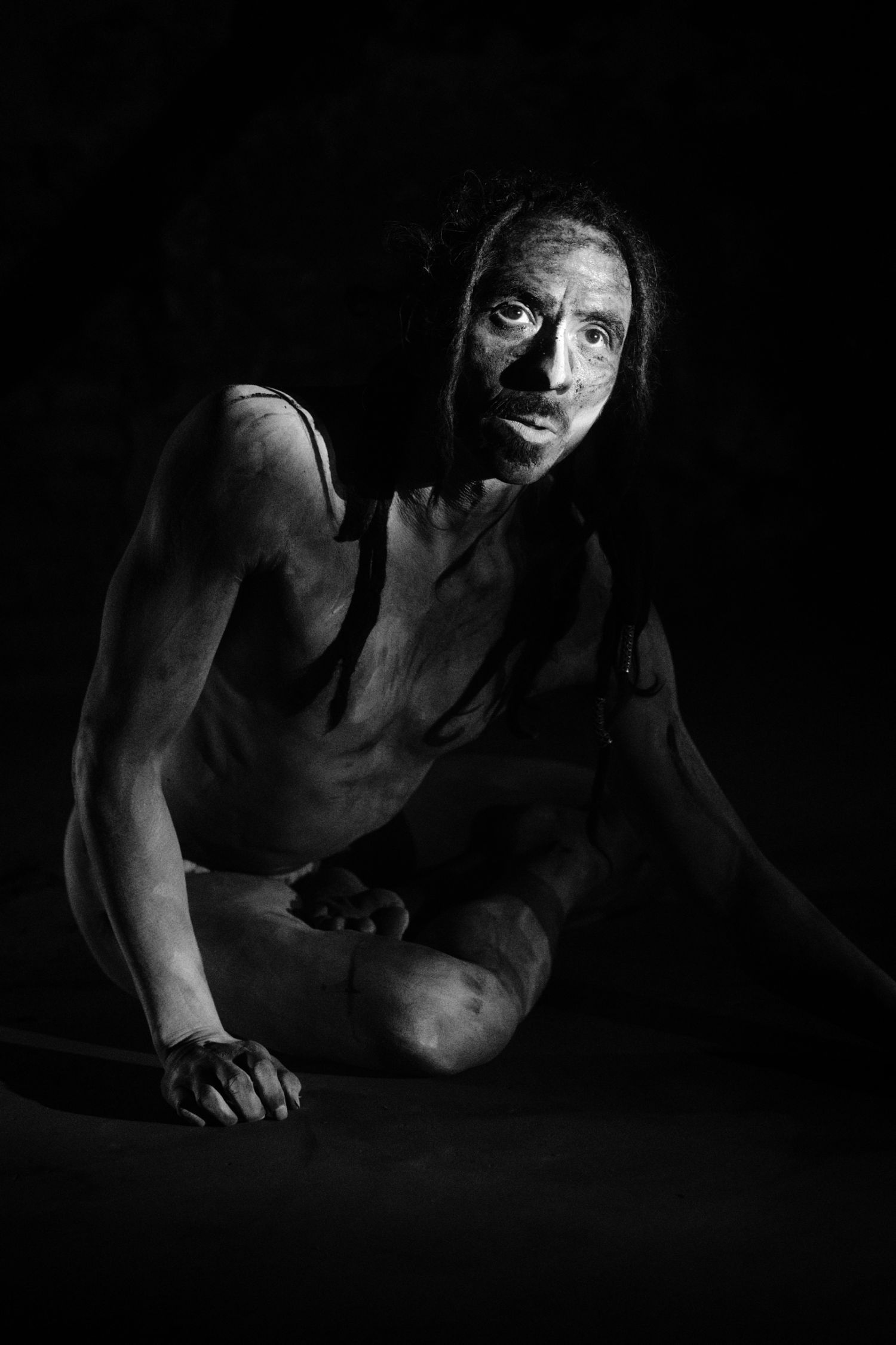 Black and white portrait of a person with dark makeup sitting on floor against dark background.