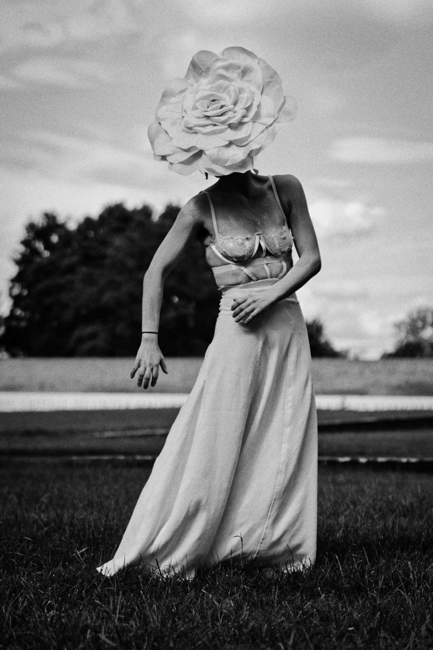 Black and white photo of a woman in a lace bralette and long skirt, her head hidden by a giant rose, standing on a grassy field.