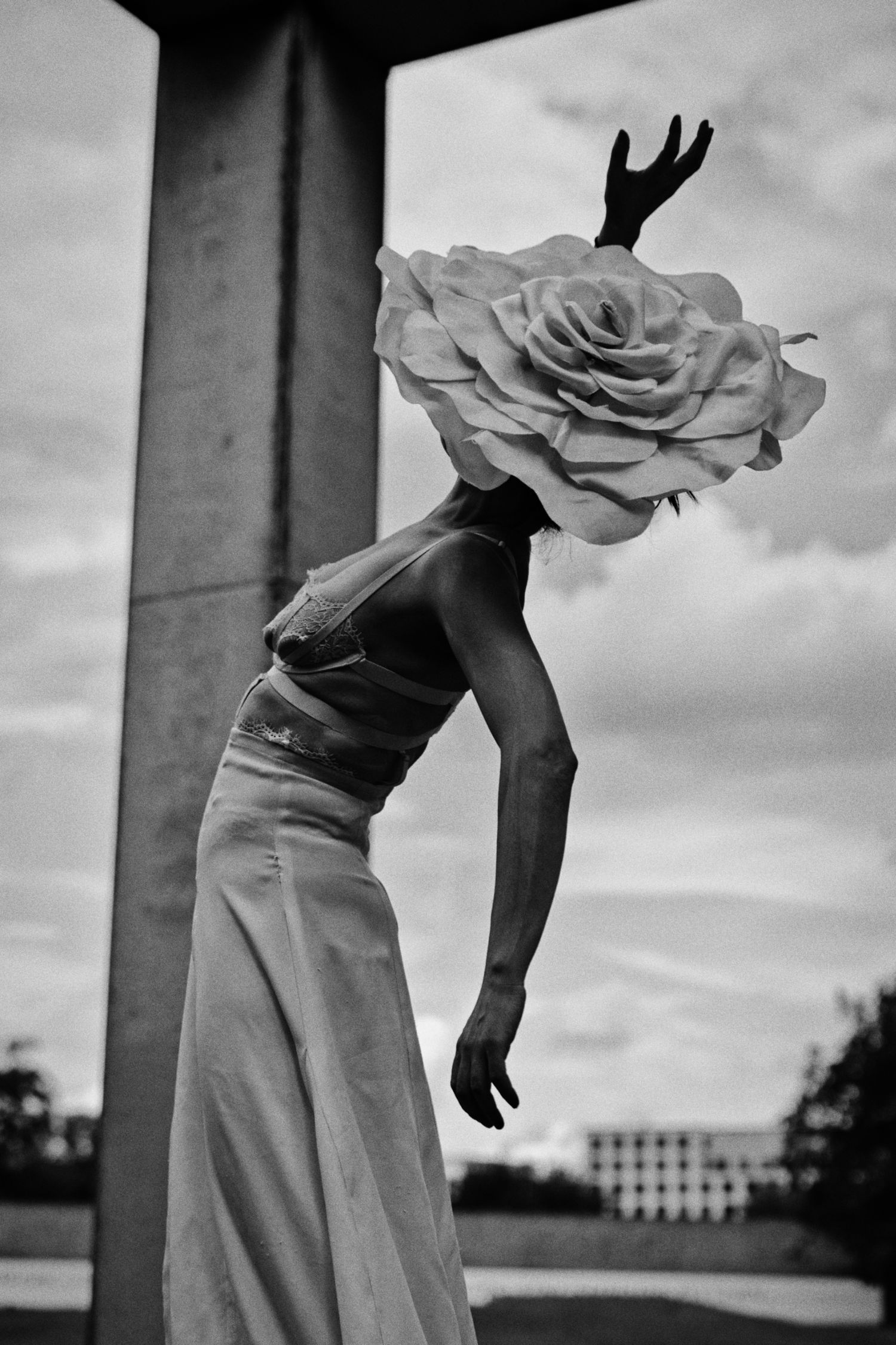 Black and white photo of a woman in a long dress leaning forward with an oversized rose covering her head beside a concrete pillar.