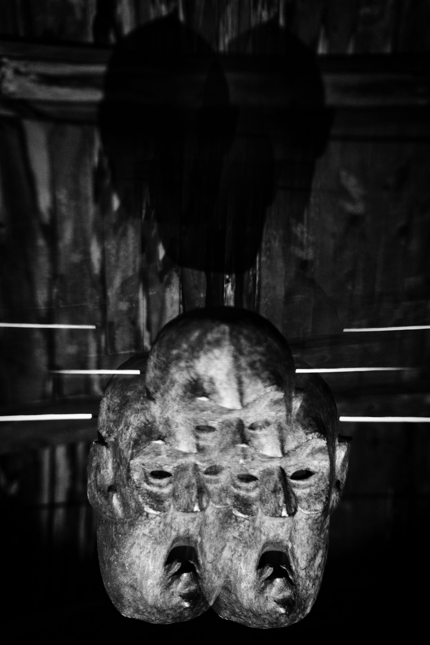 Black and white photo of an ancient wooden tribal mask with multiple faces, hanging dramatically in dark surroundings.