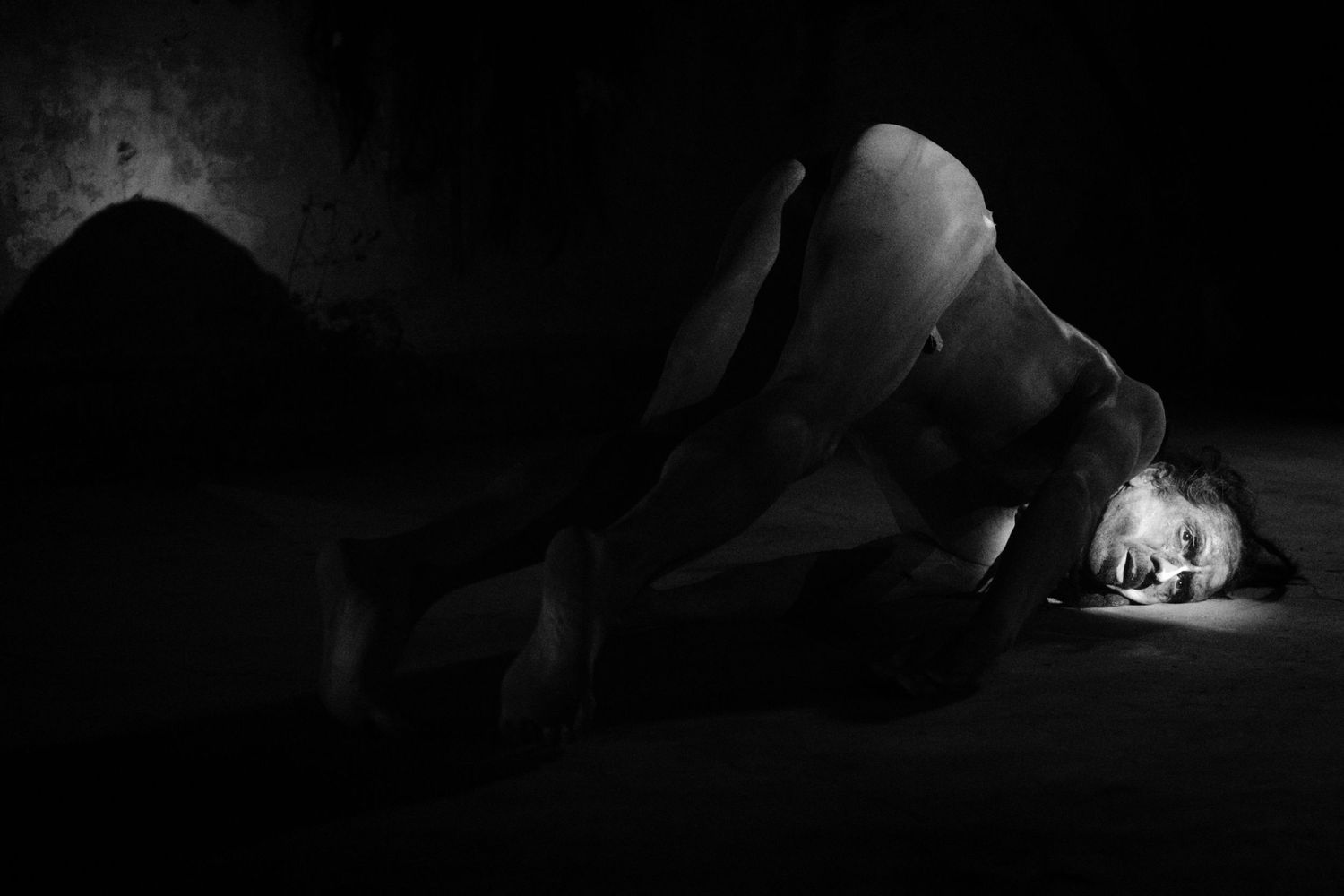 Black and white photo of a person curled up on a dark floor, illuminated by soft light.