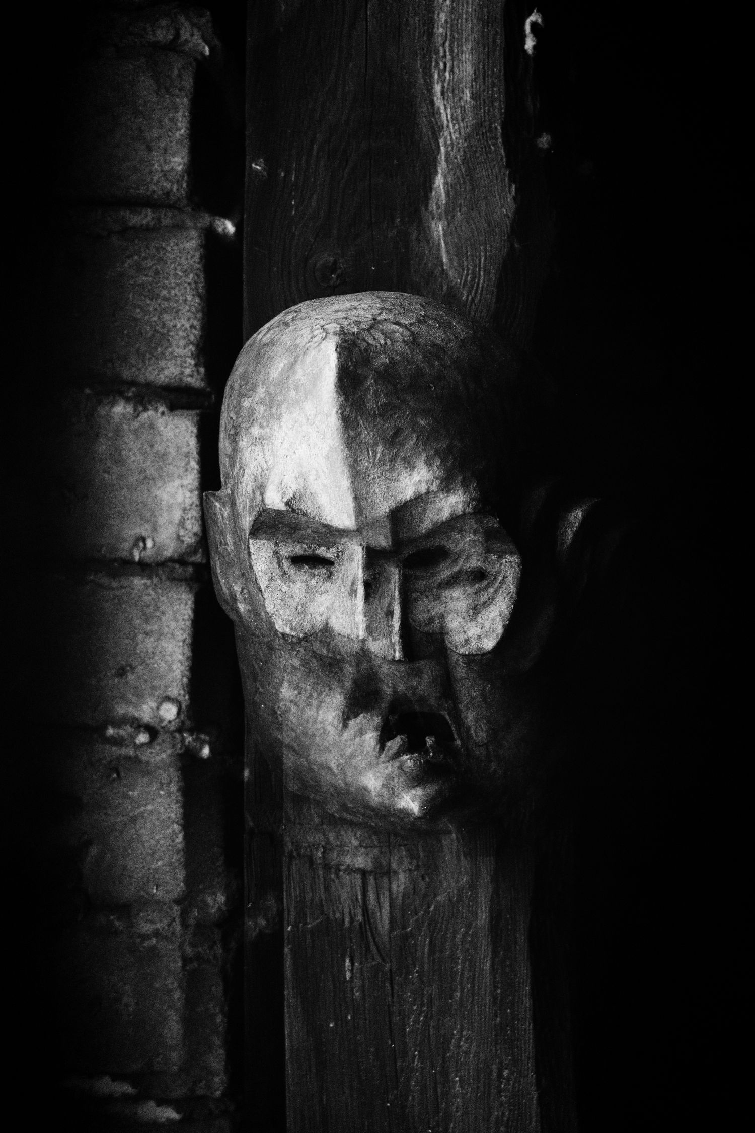 Black and white photo of a carved wooden face mask peeking from behind a dark wooden post.