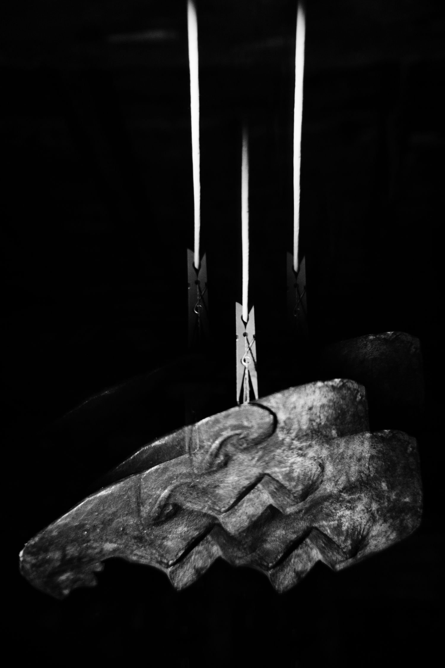 Black and white photo of a carved stone artifact suspended by a clothespin on white strings against dark background.