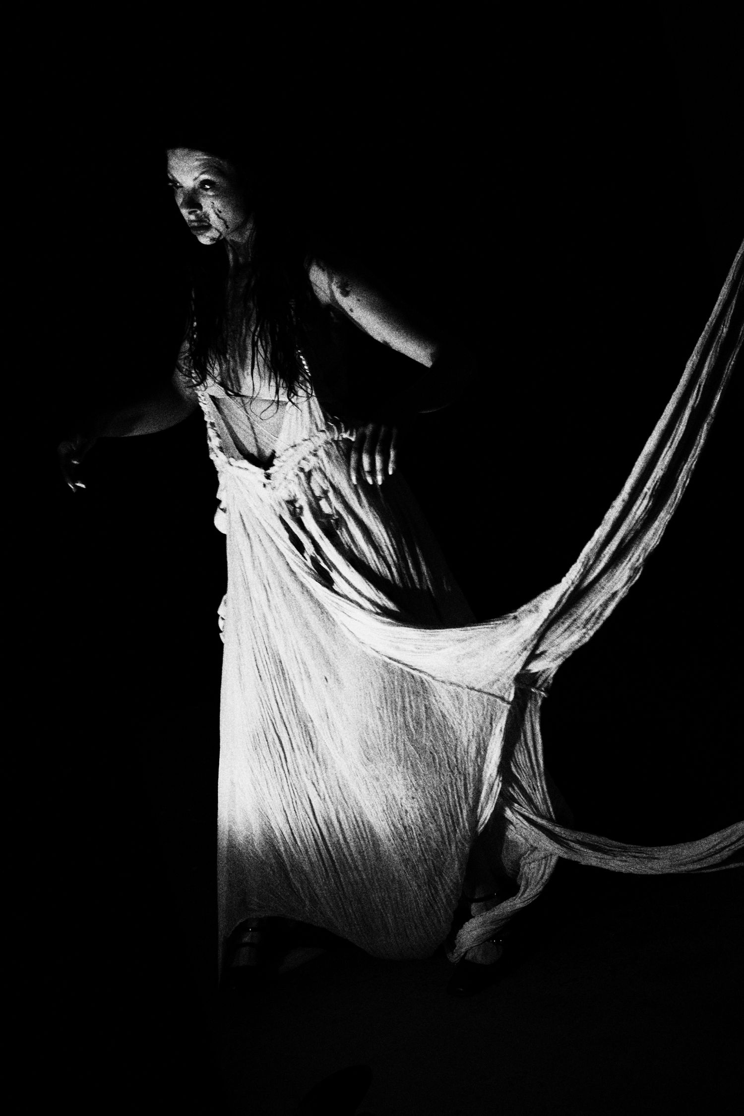 Black and white photo of a woman in a flowing white dress dancing dramatically against a dark background.