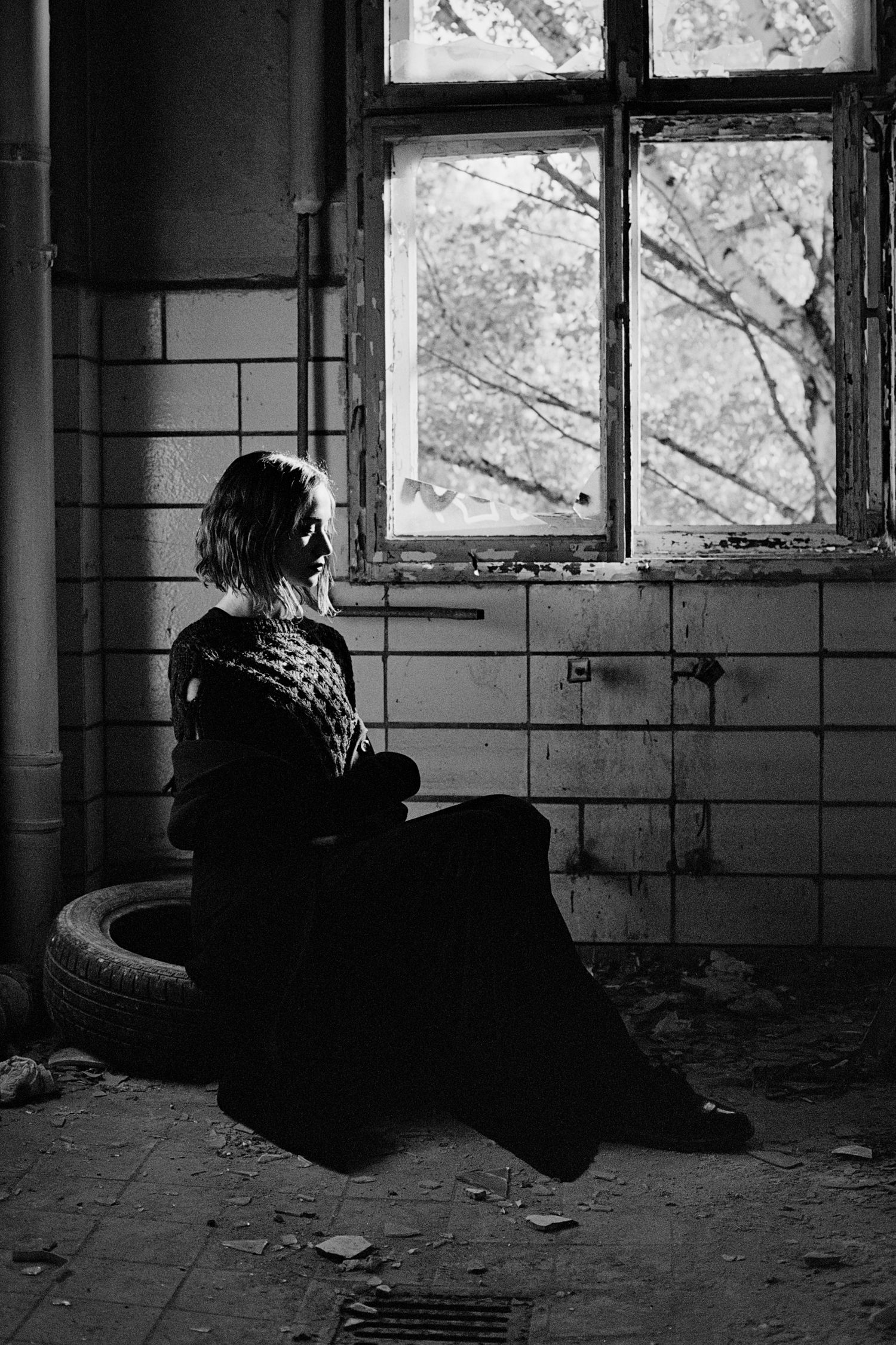 Black and white photo of a woman sitting on a tire in an abandoned building, gazing through a broken window.