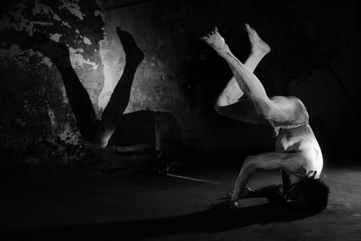 Black and white photo of a dancer performing an acrobatic pose on a dark stage with dramatic shadows.