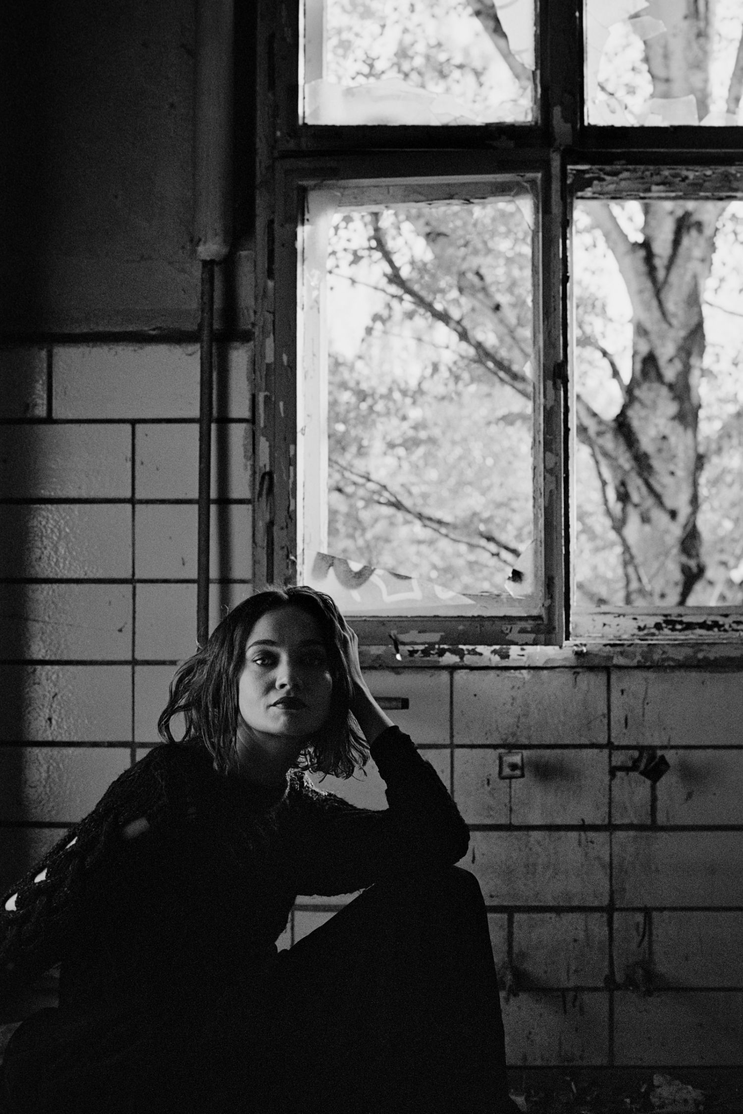 Black and white photo of a young woman sitting in an abandoned building near a weathered window with trees visible outside.