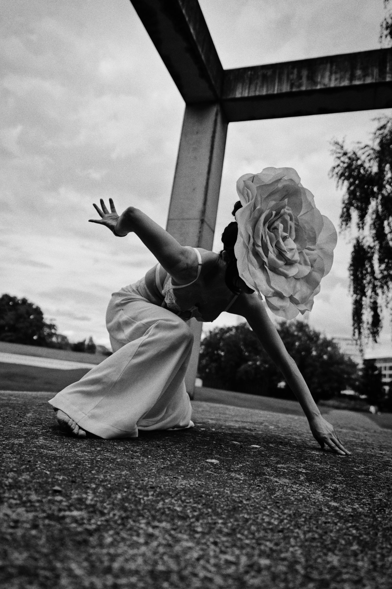Black and white photo of a dancer in a crouched pose with a giant rose covering her head, beneath a large geometric steel structure.