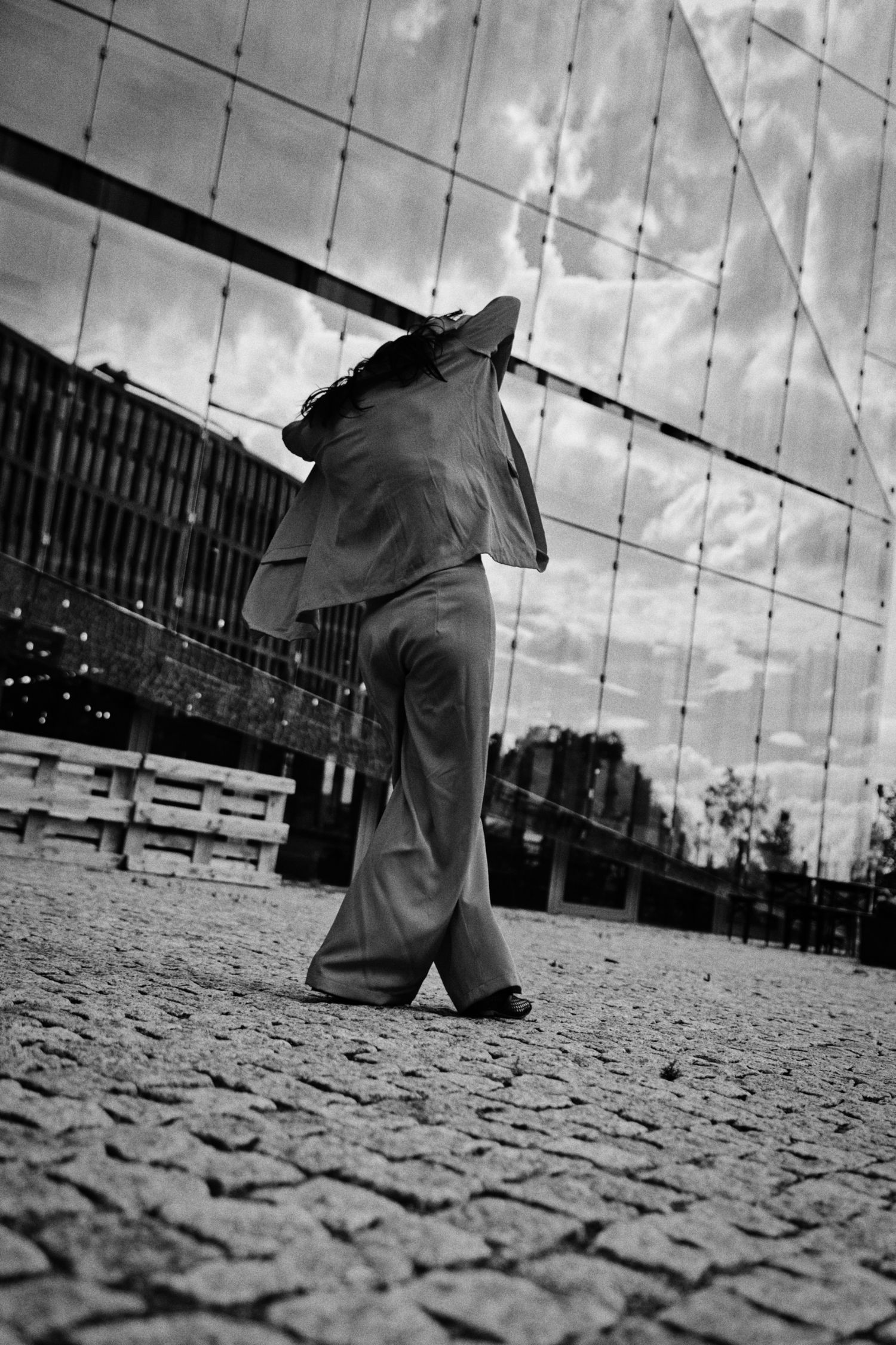 Black and white photo of a person in a loose suit and wide-leg trousers standing on cobblestones before a mirrored wall reflecting the sky.