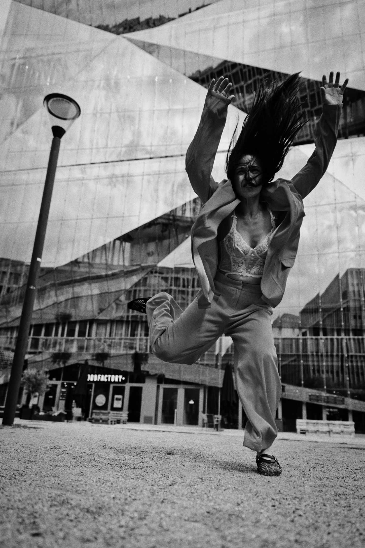 Black and white photo of a woman in a loose suit leaping mid-air, hair flying, in front of a faceted glass building and street lamp.