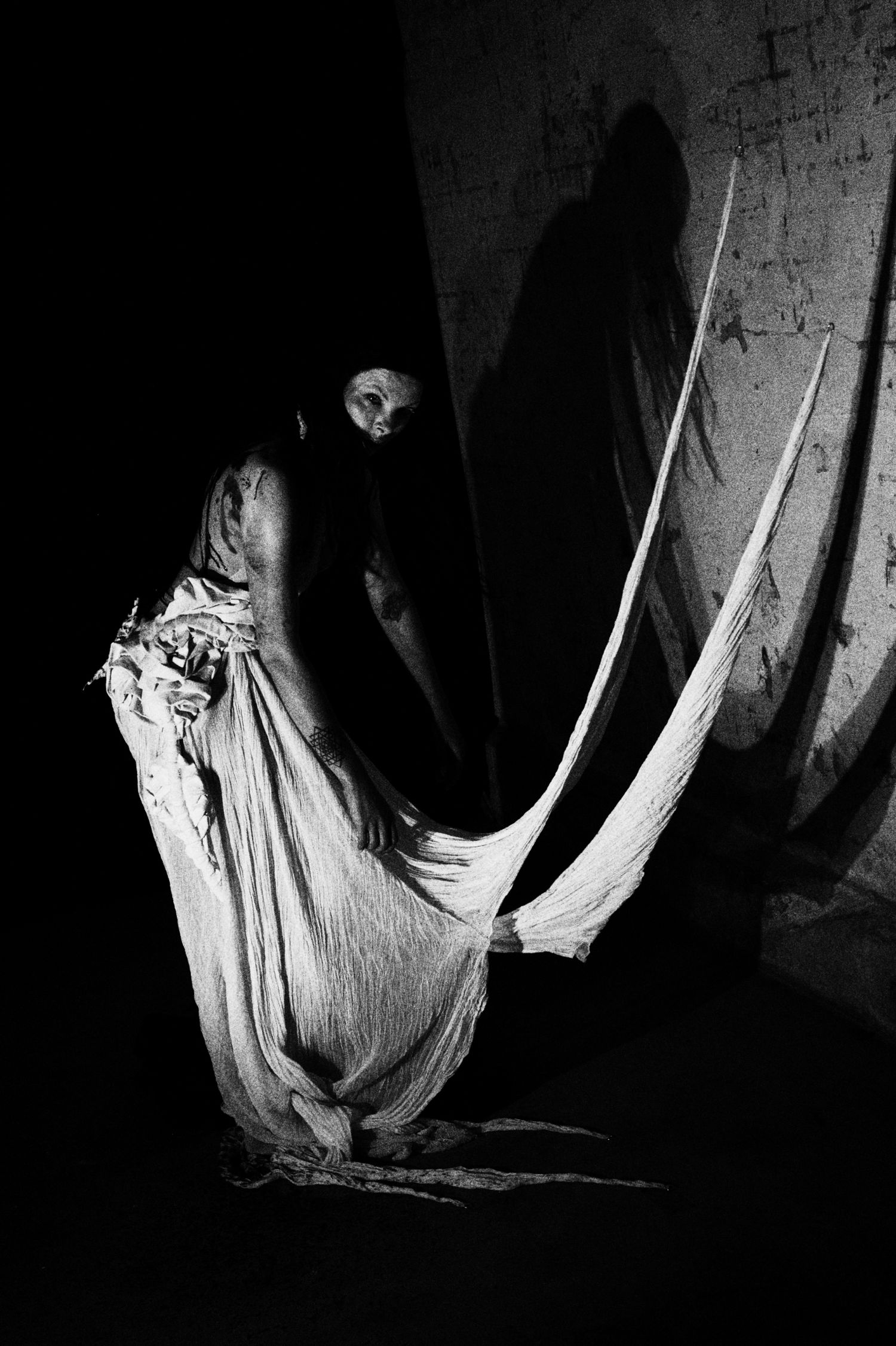 Dramatic black and white photo of a figure in tattered white robes holding fabric, casting an eerie shadow on a dark wall.
