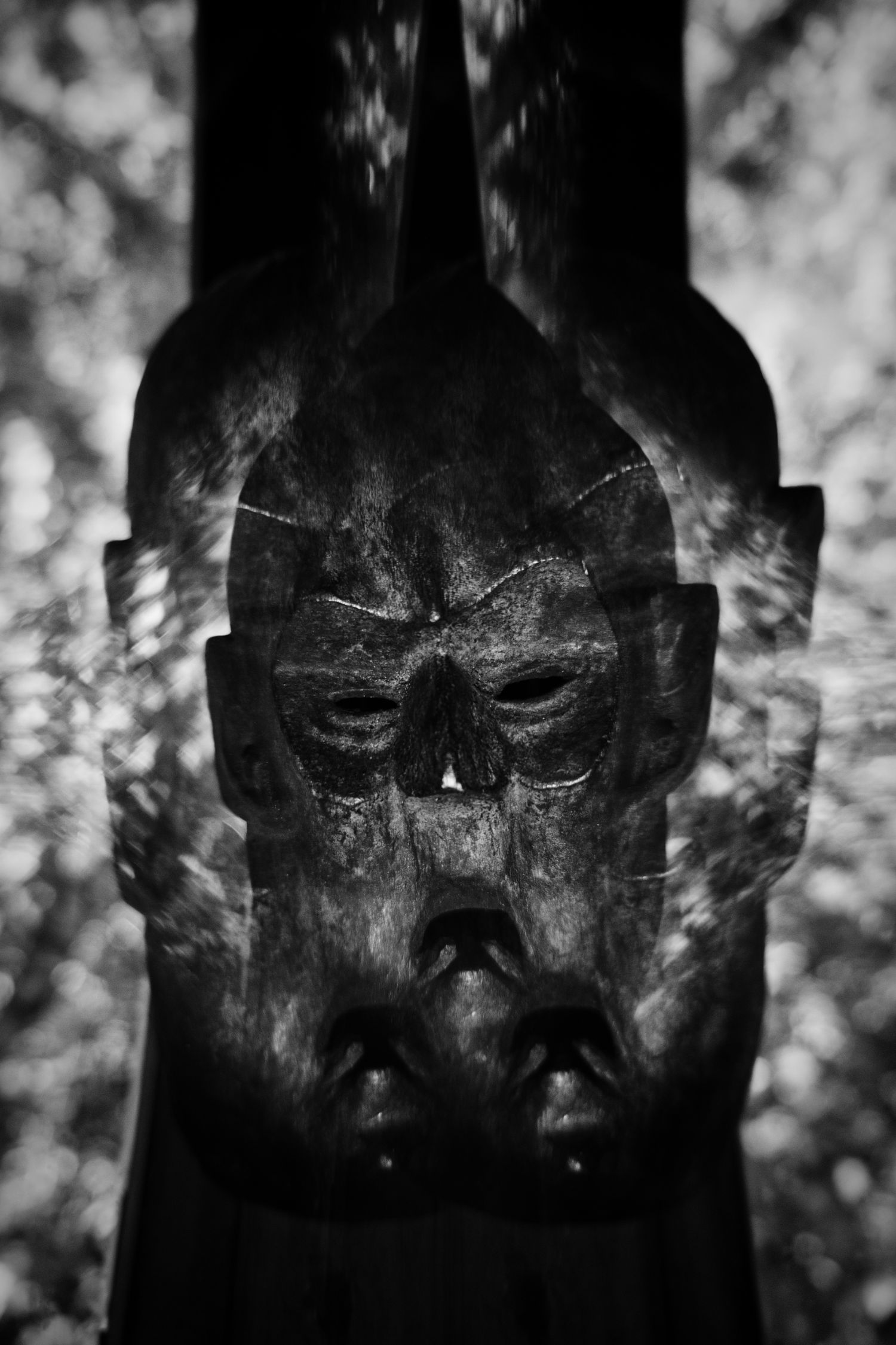 Dark black and white double exposure of an ancient tribal wooden mask with haunting hollow eyes and open mouth.
