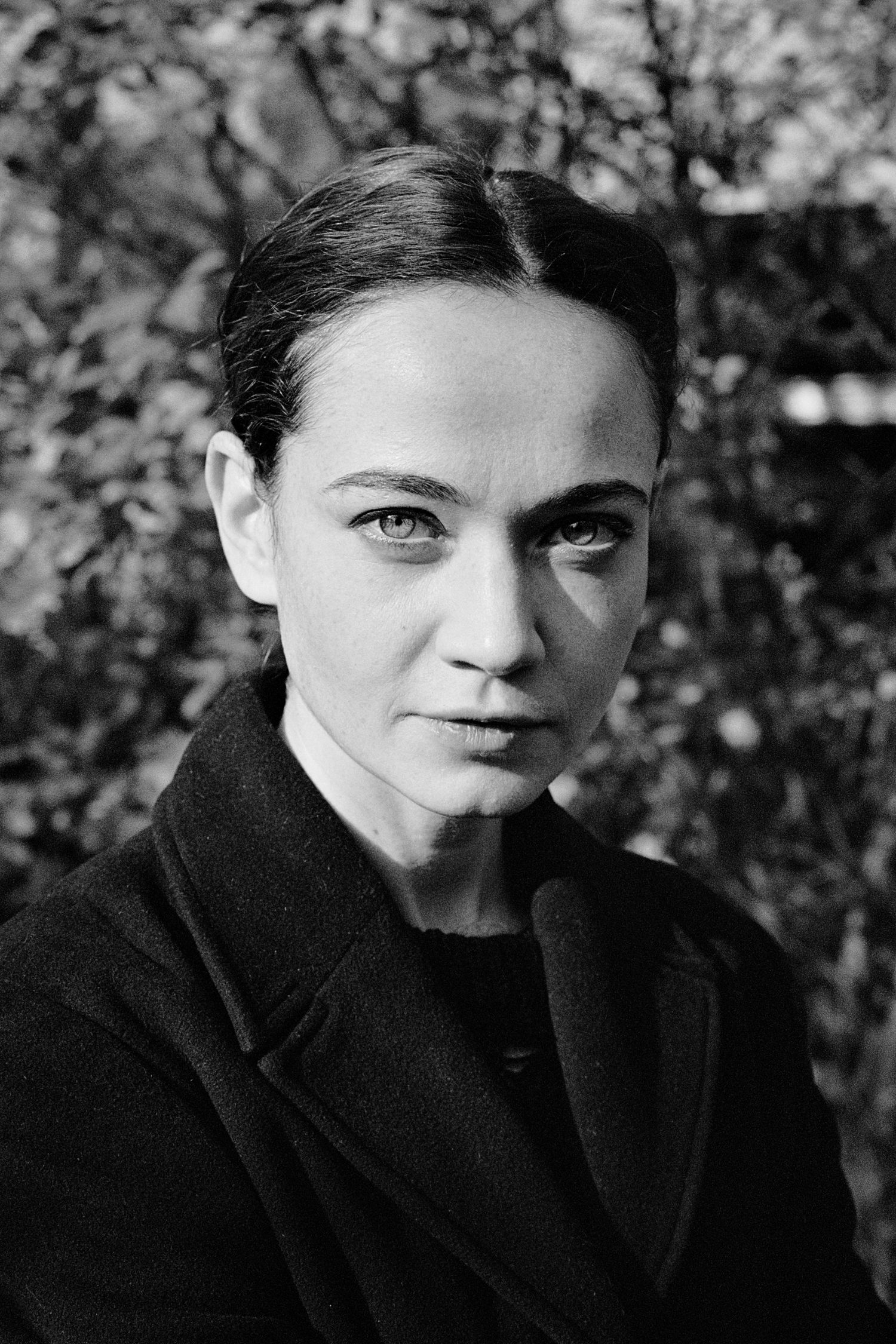 Black and white portrait of a young woman with slicked-back hair wearing a dark coat, set against a leafy background.