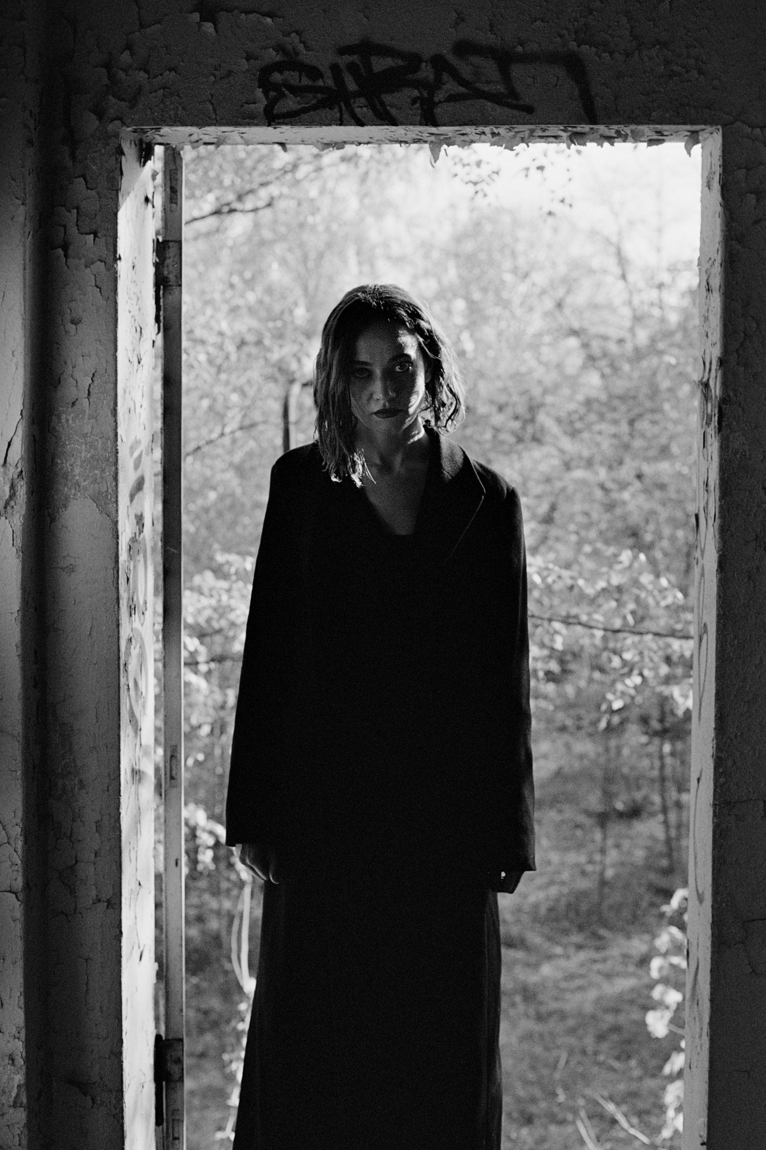 Black and white photo of a woman in dark dress standing in abandoned doorway with graffiti and overgrown trees behind her.