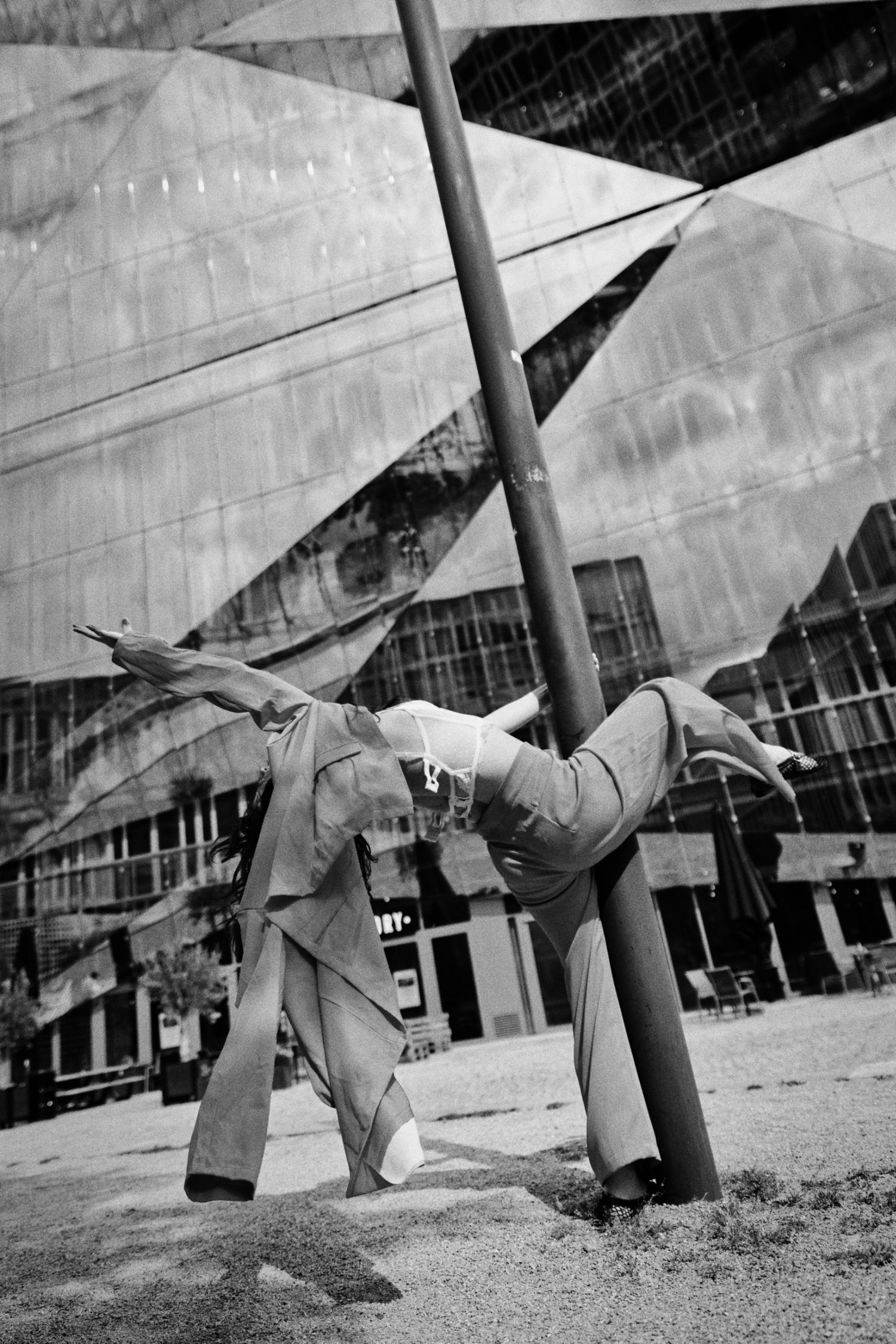 Black and white photo of a person in a suit performing a handstand split against a lamppost in front of a mirrored glass building.