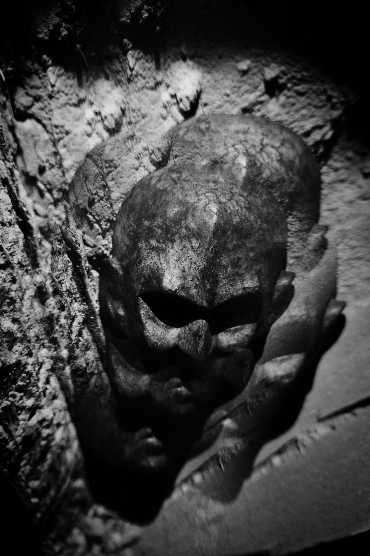Dark black and white photo of an ancient cracked skull sculpture emerging from a rough stone wall.