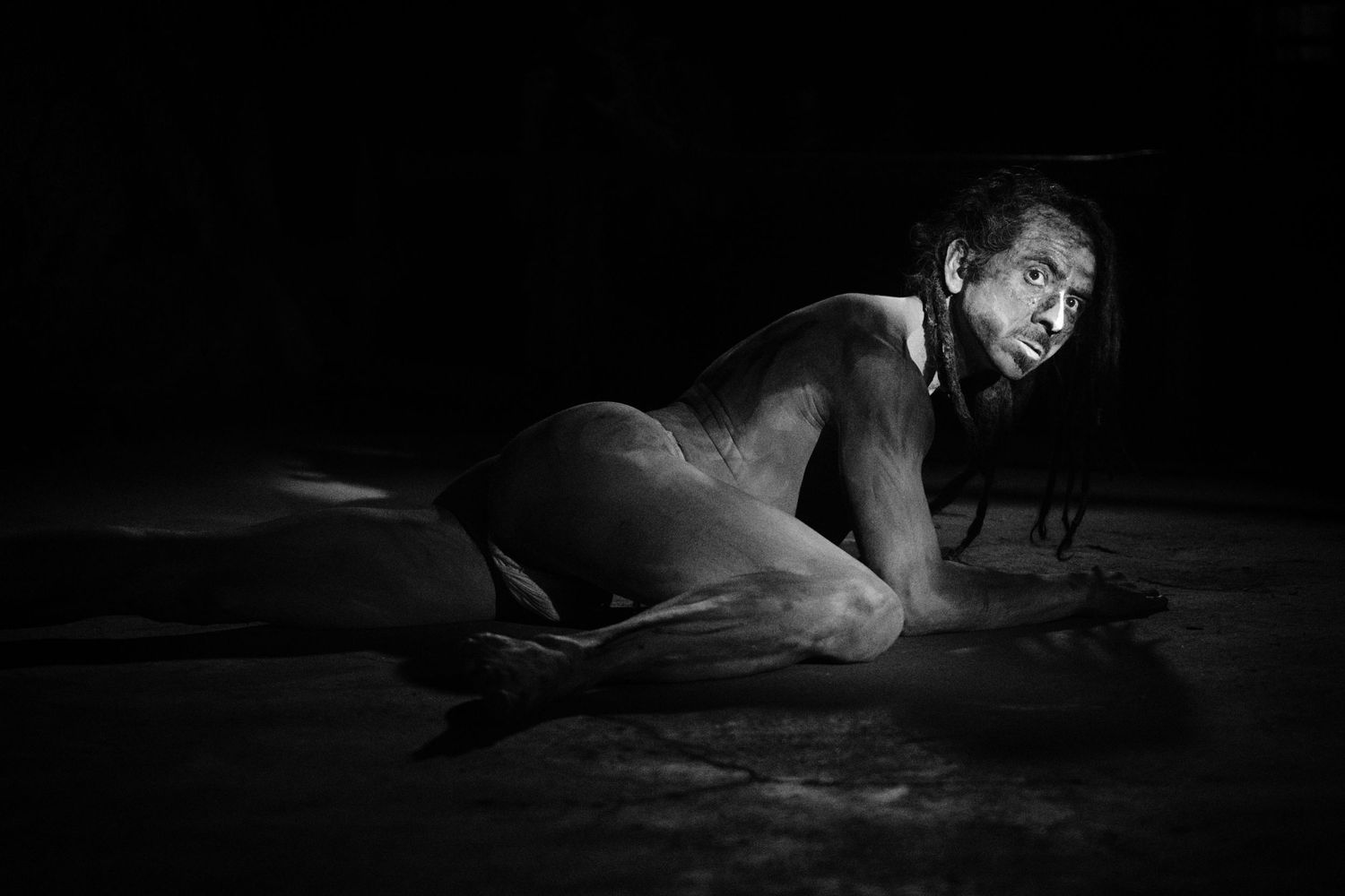 Black and white photo of a shirtless person with dreadlocks crawling on a dark wet floor dramatically.