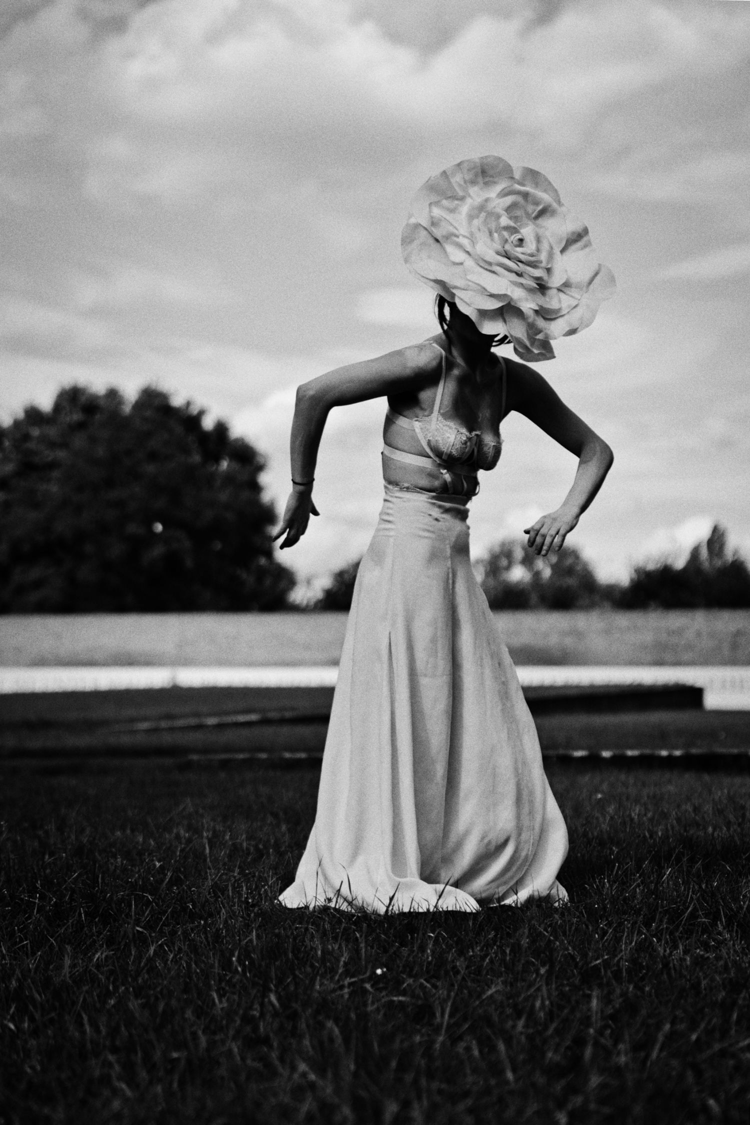 Black and white photo of a woman in a flowing long skirt and crop top dancing on a grassy field, wearing a giant flower headpiece.
