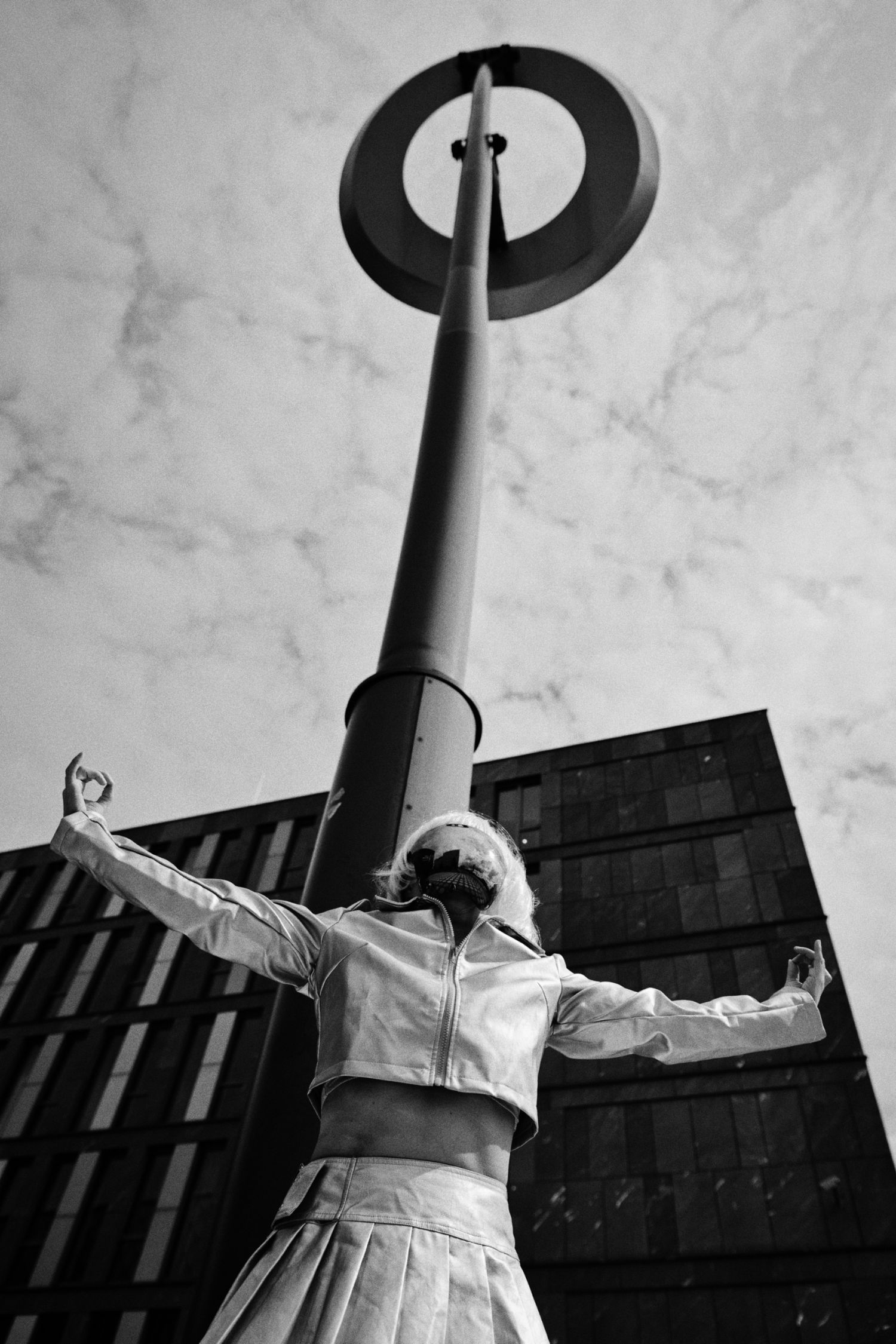 Black and white low-angle photo of a woman in a cropped jacket and pleated skirt with arms outstretched beside a tall ring-topped lamppost.