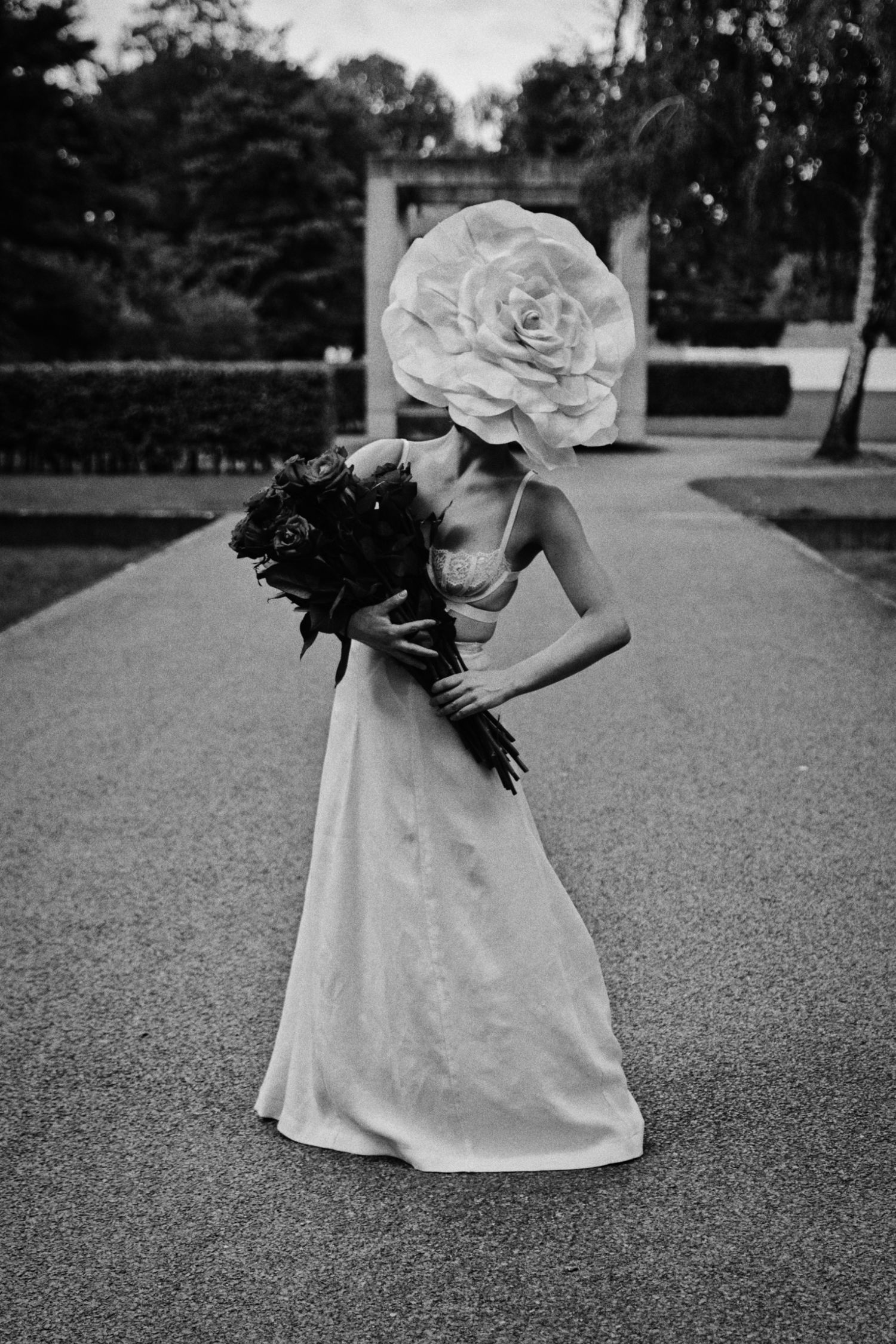 Black and white photo of a woman in a satin gown holding a dark bouquet, her face hidden by an oversized paper flower, in a park.