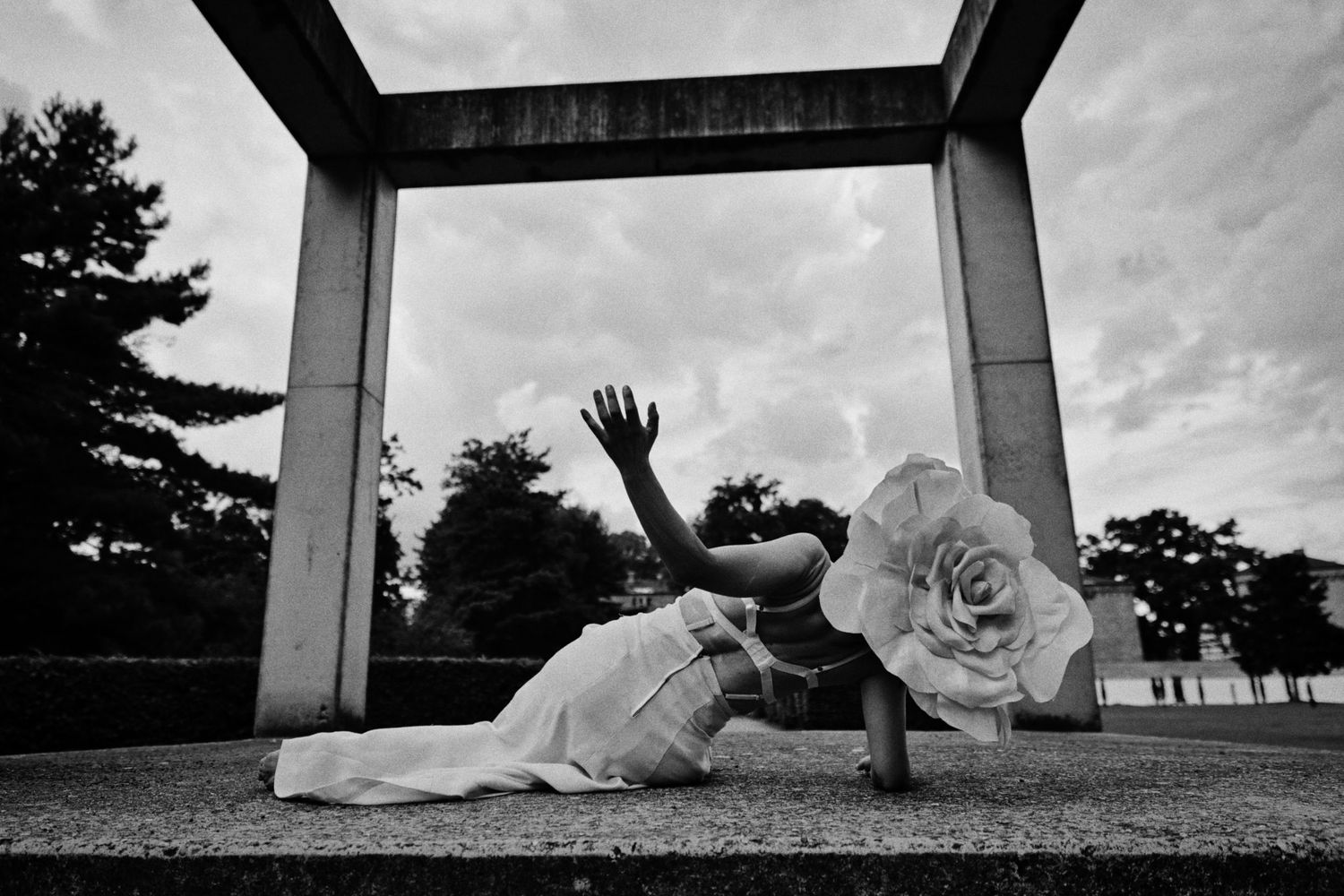 Black and white photo of a dancer in a white dress reaching upward on a stone platform beneath a concrete frame, with a large paper flower.
