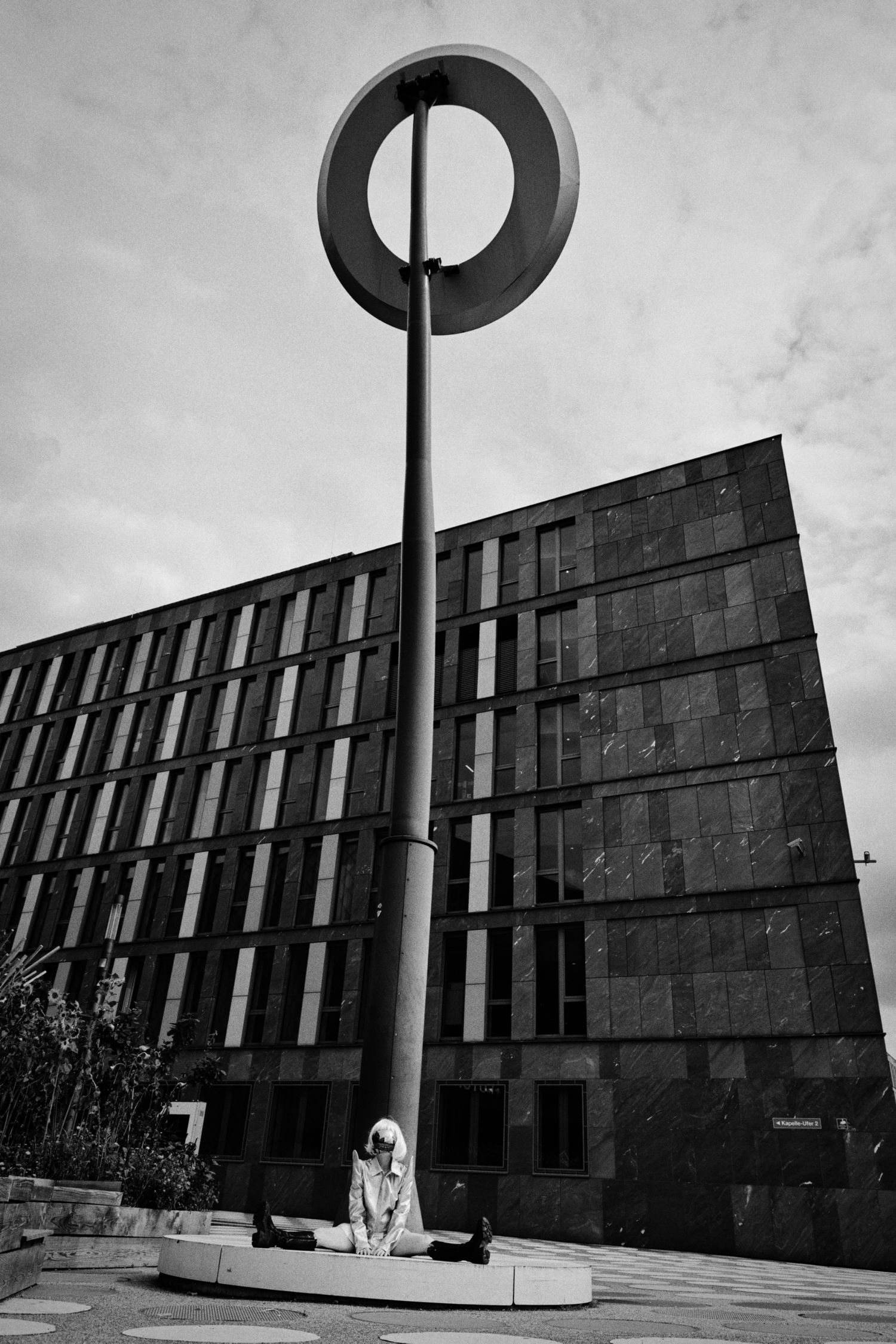 Black and white photo of a person sitting on a circular bench beneath a tall ring-topped pole in front of a modern building.