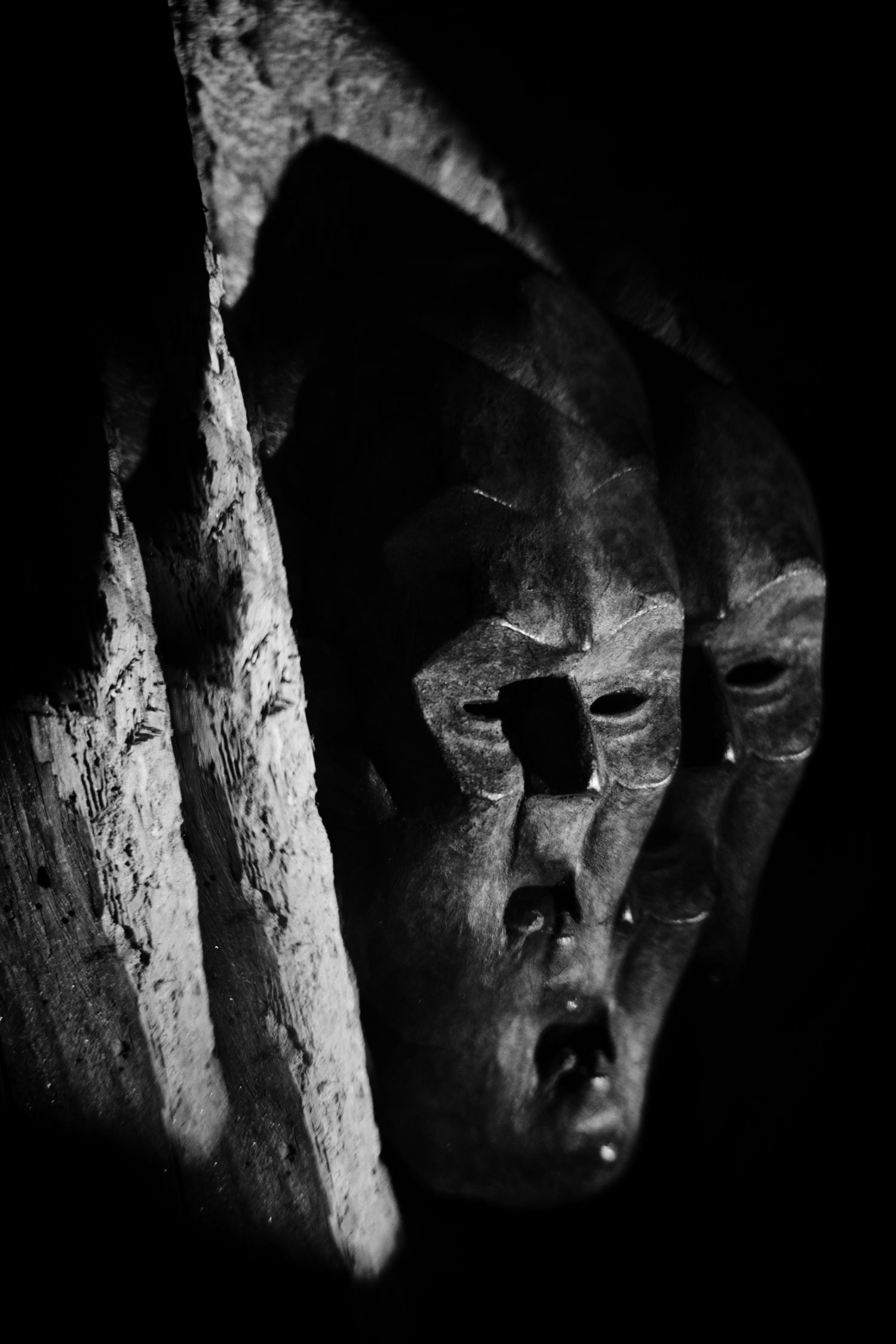 Black and white photo of a carved wooden African tribal mask partially obscured by shadow and splintered wood.