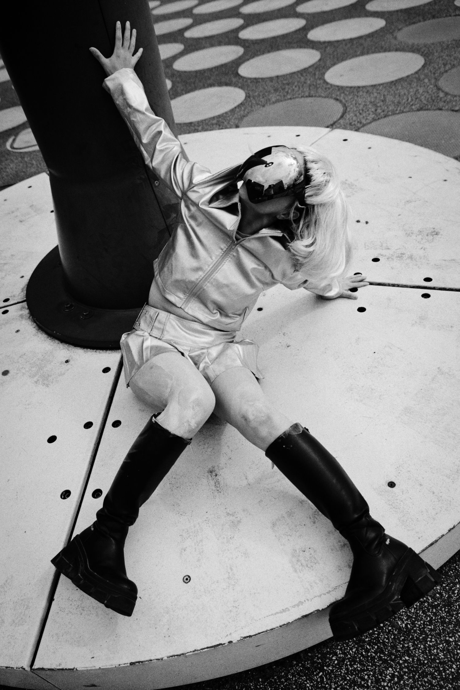 Black and white photo of a person in a shiny jacket, shorts and tall black boots reclining against a dark pole on a large circular platform.