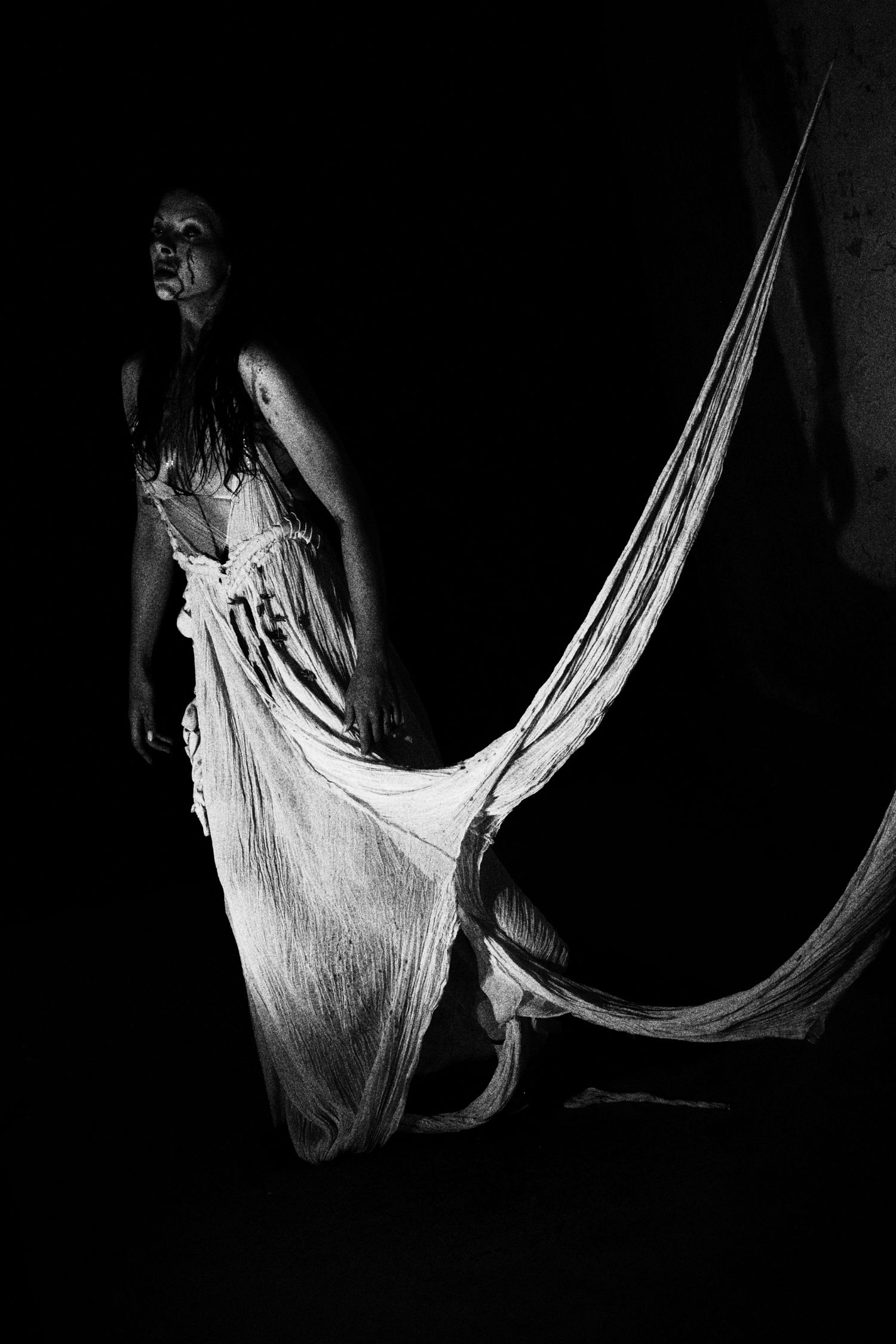 Black and white photo of a woman in a flowing white gown standing dramatically in darkness.