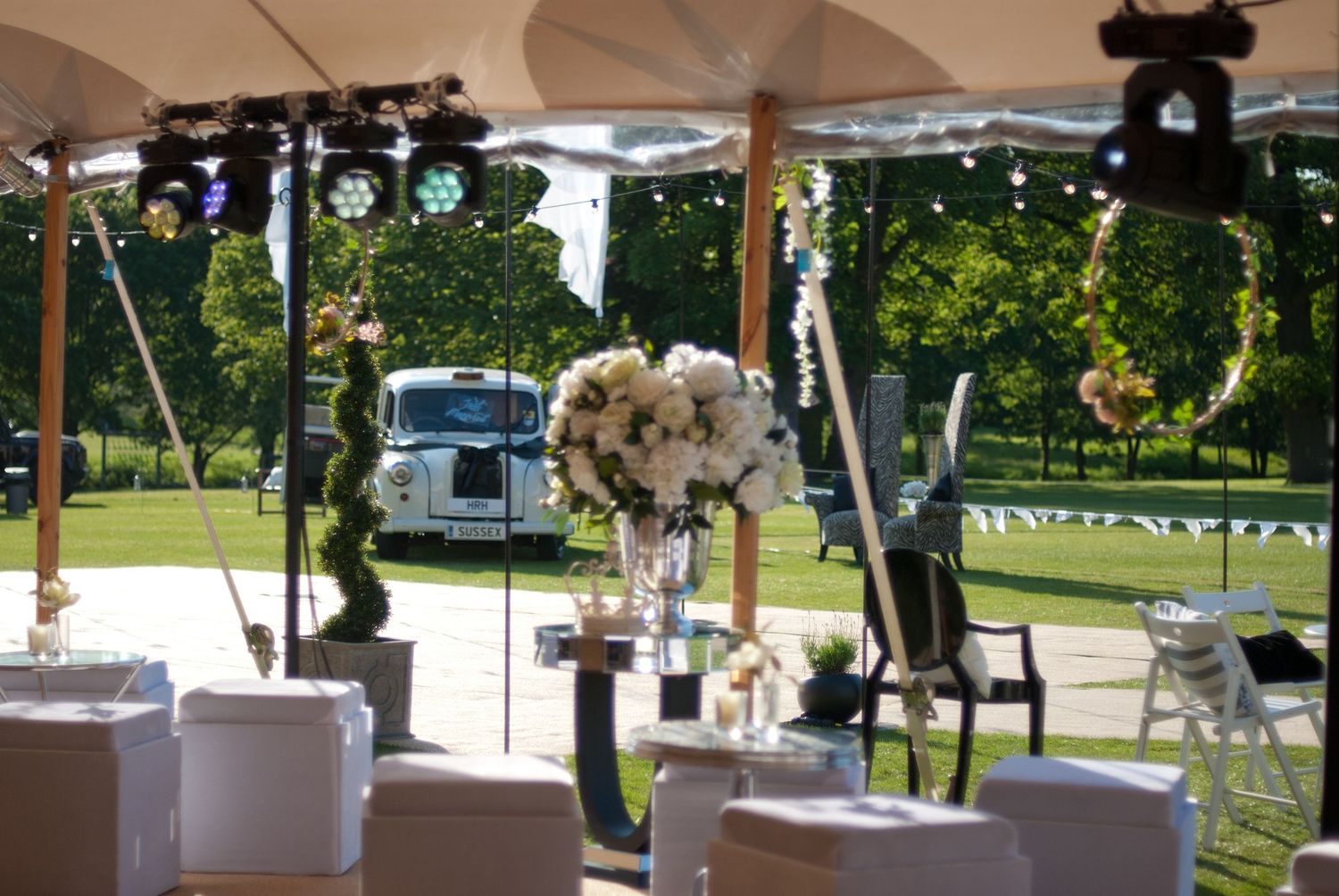 Elegant outdoor wedding venue under a white tent with floral arrangements, fairy lights, and a vintage white car.