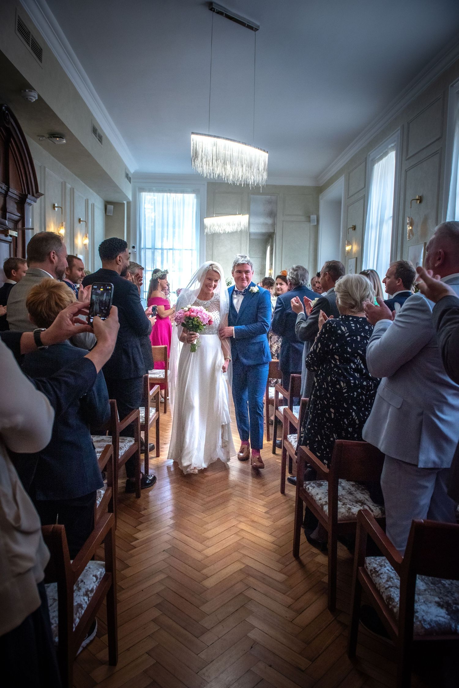 Bride and groom walk down the aisle as guests applaud in an elegant ceremony room. Chelsea Town Hall Wedding, London.