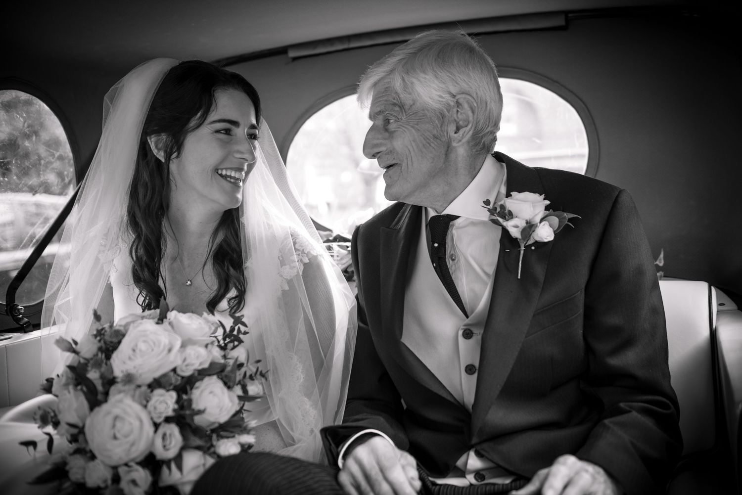 Bride and father sharing a smile in a vintage wedding car, black and white.