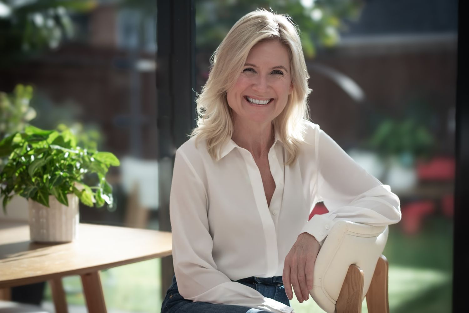 Cheerful blonde woman in white blouse smiling while seated at a wooden table on a sunny patio.