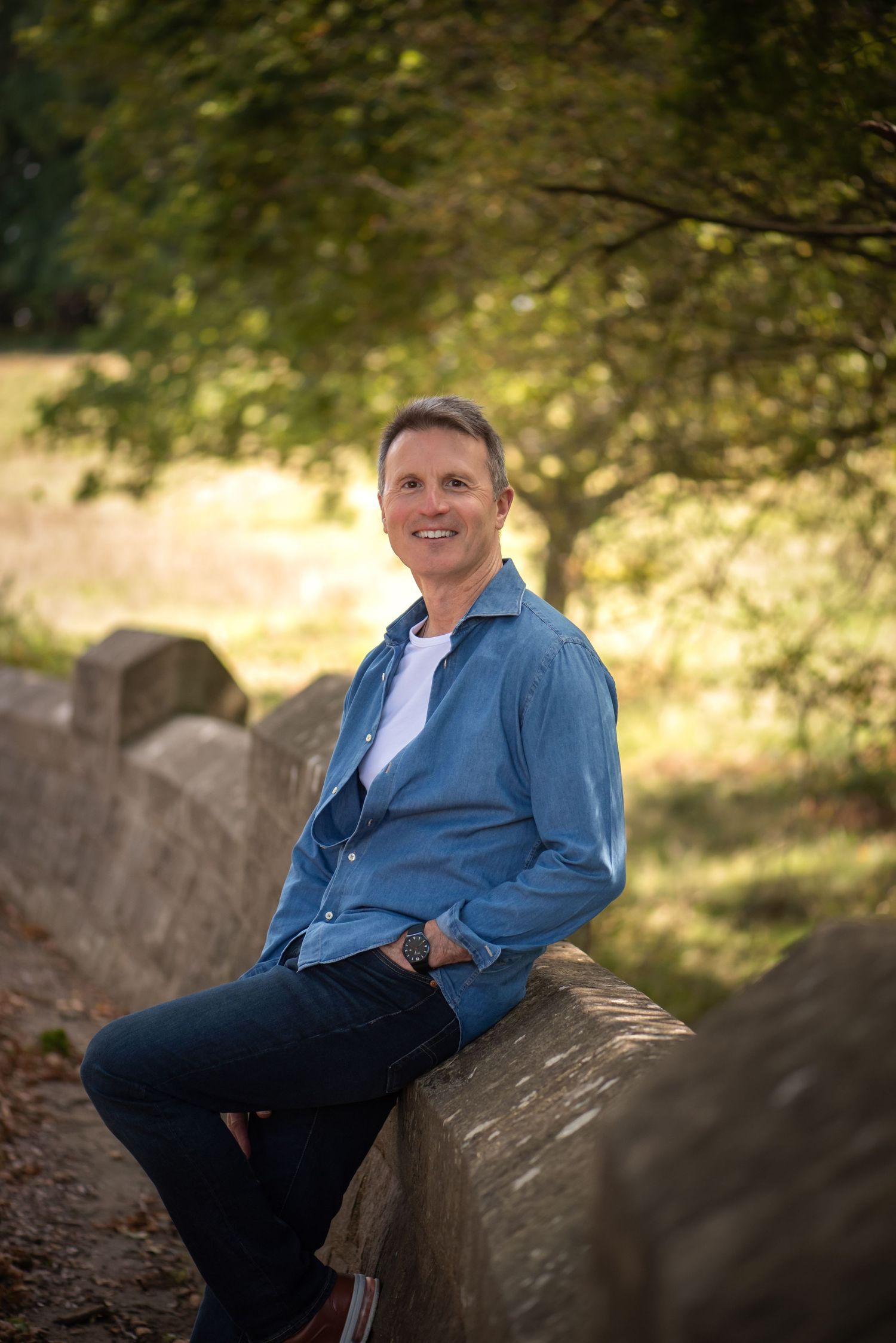 Smiling middle-aged man in blue denim shirt sitting on a stone wall in nature. business portrait