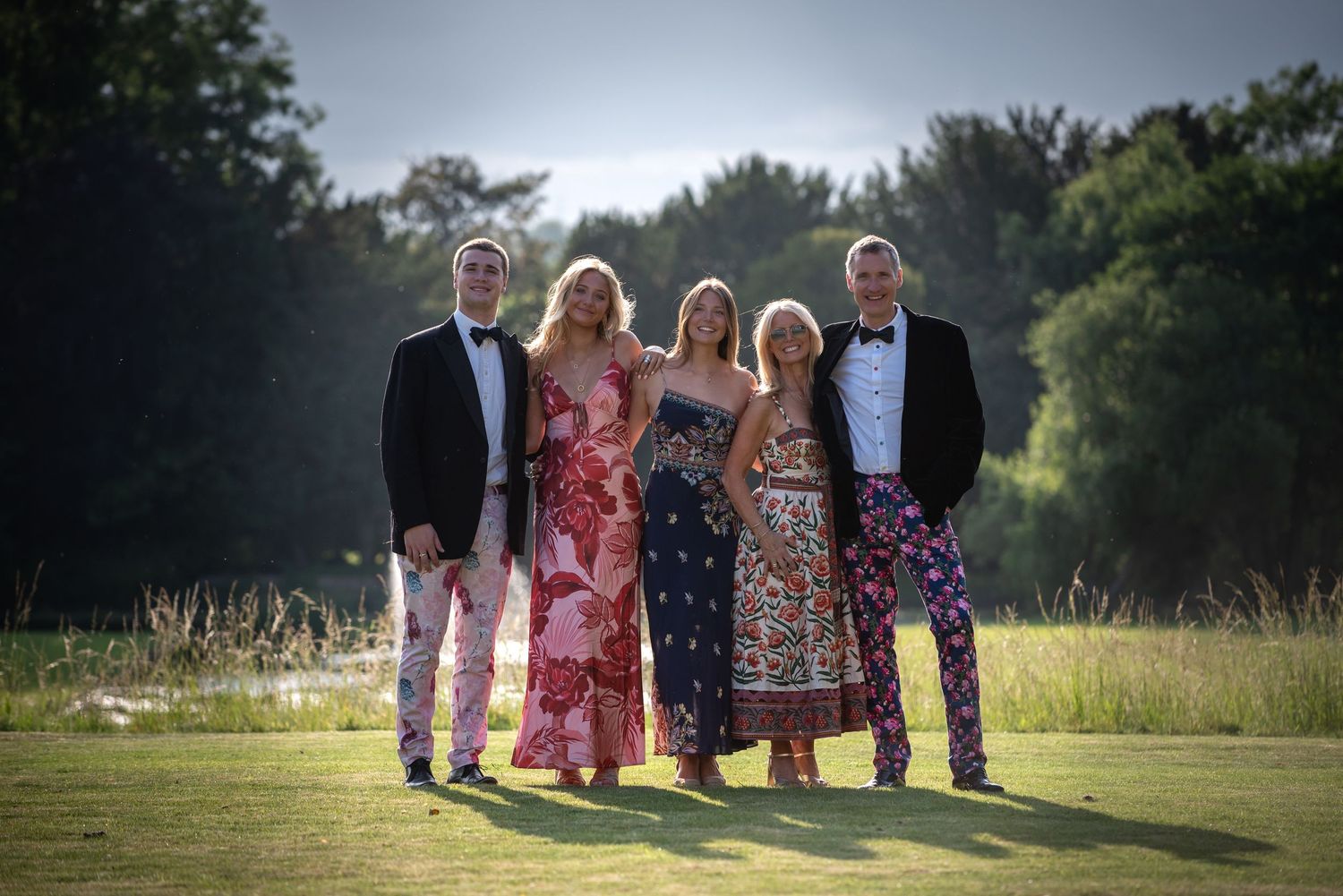 Five elegantly dressed guests in colorful formal wear posing together on a sunlit garden lawn.