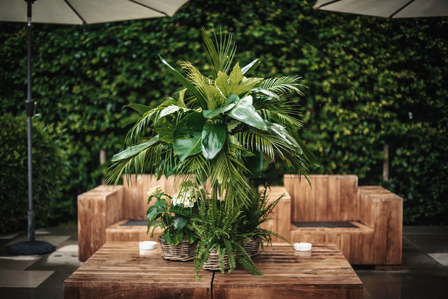 Tropical palm leaf centerpiece on wooden table with candles against a green hedge backdrop.