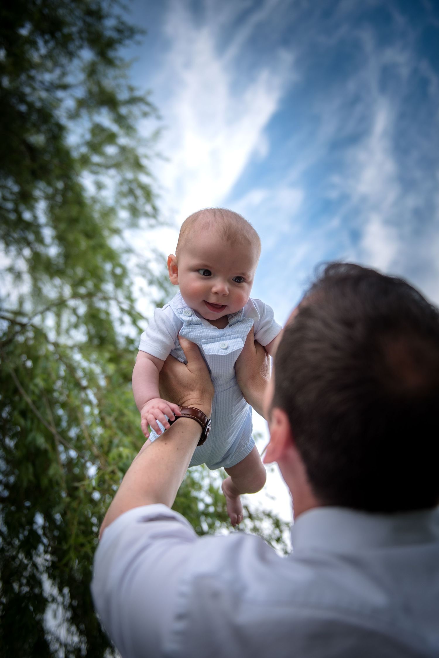 Father lifting happy baby up against a blue sky with trees.