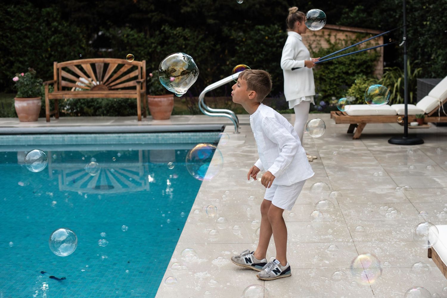 Two children in white outfits playing with bubbles beside a bright blue swimming pool.