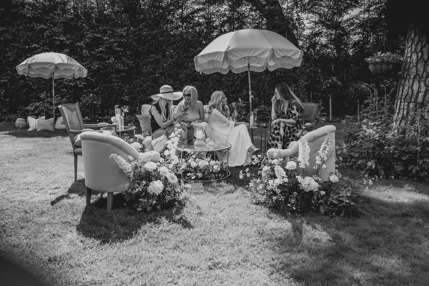 Black and white outdoor garden party scene with guests seated under parasols surrounded by floral arrangements.